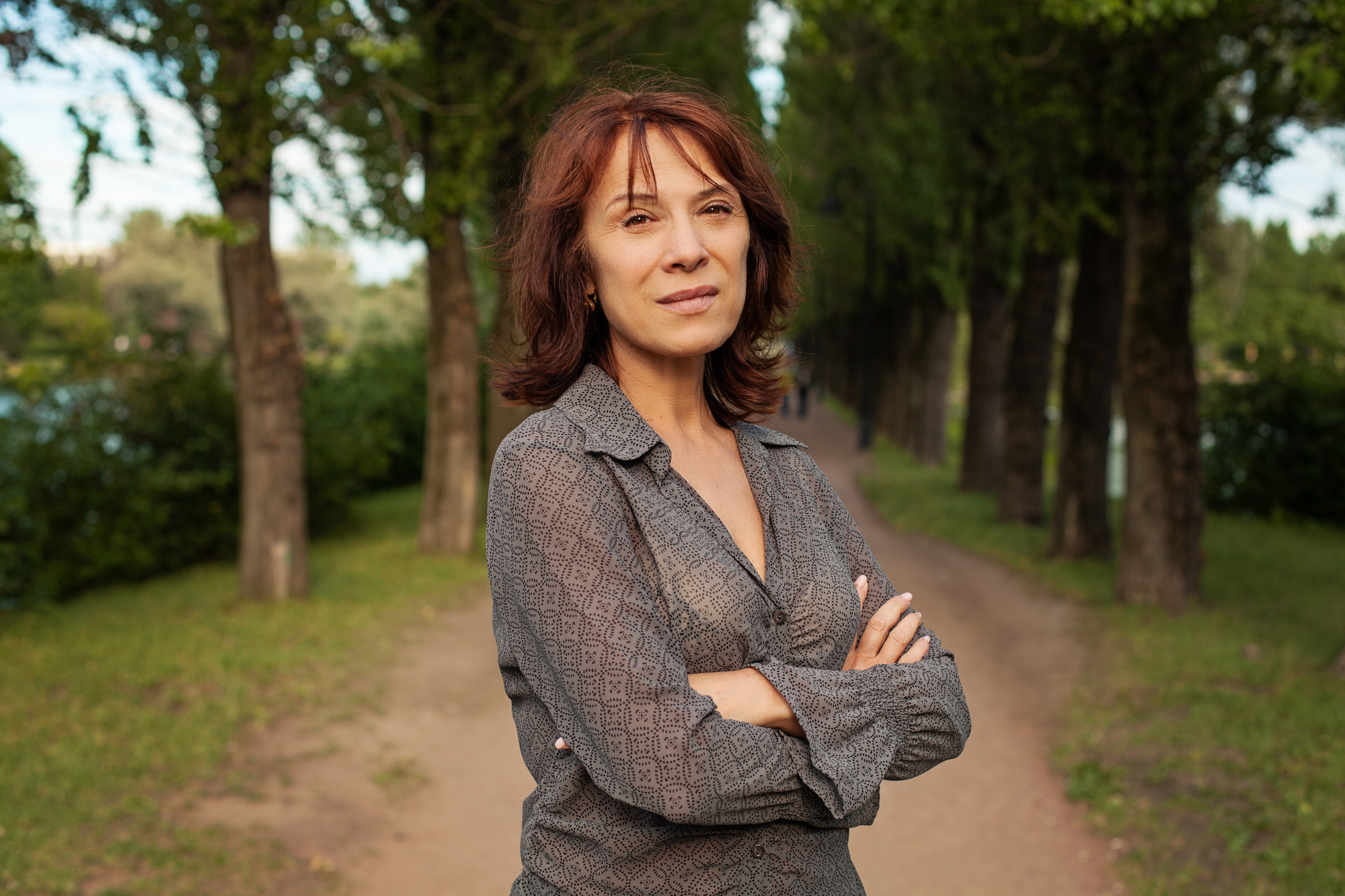 A woman with shoulder-length brown hair stands with her arms crossed on a tree-lined path. She is wearing a patterned gray blouse and looks directly into the camera. The background features a lush, green park with trees and a clear, bright sky.