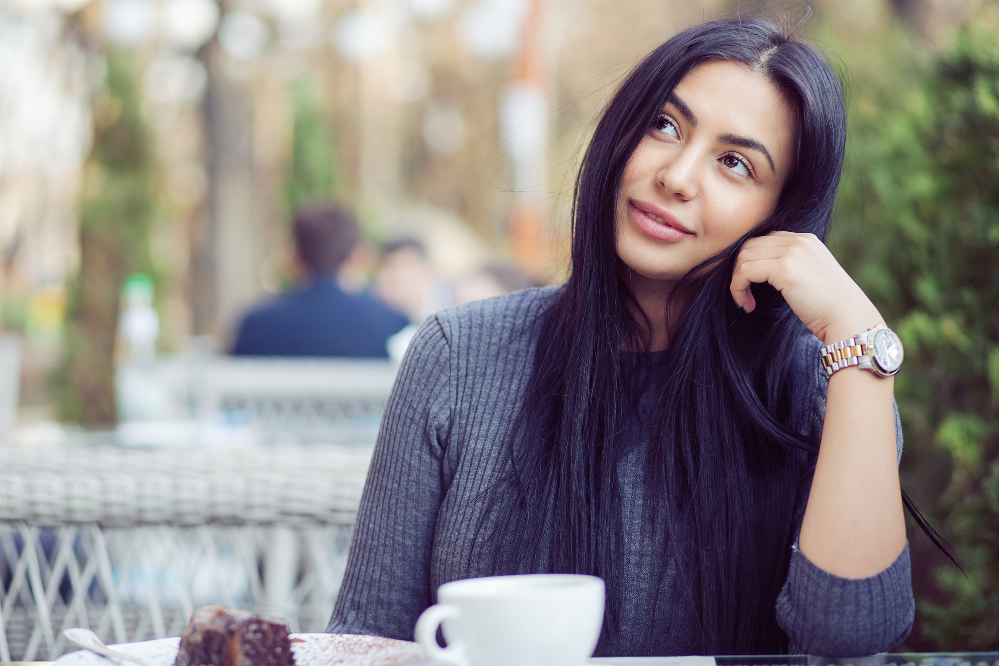 A woman with long black hair sits at an outdoor café, resting her head on her hand and smiling thoughtfully. She wears a grey sweater and a watch. In front of her is a white cup and a slice of chocolate cake. The background is blurred with greenery and other patrons.