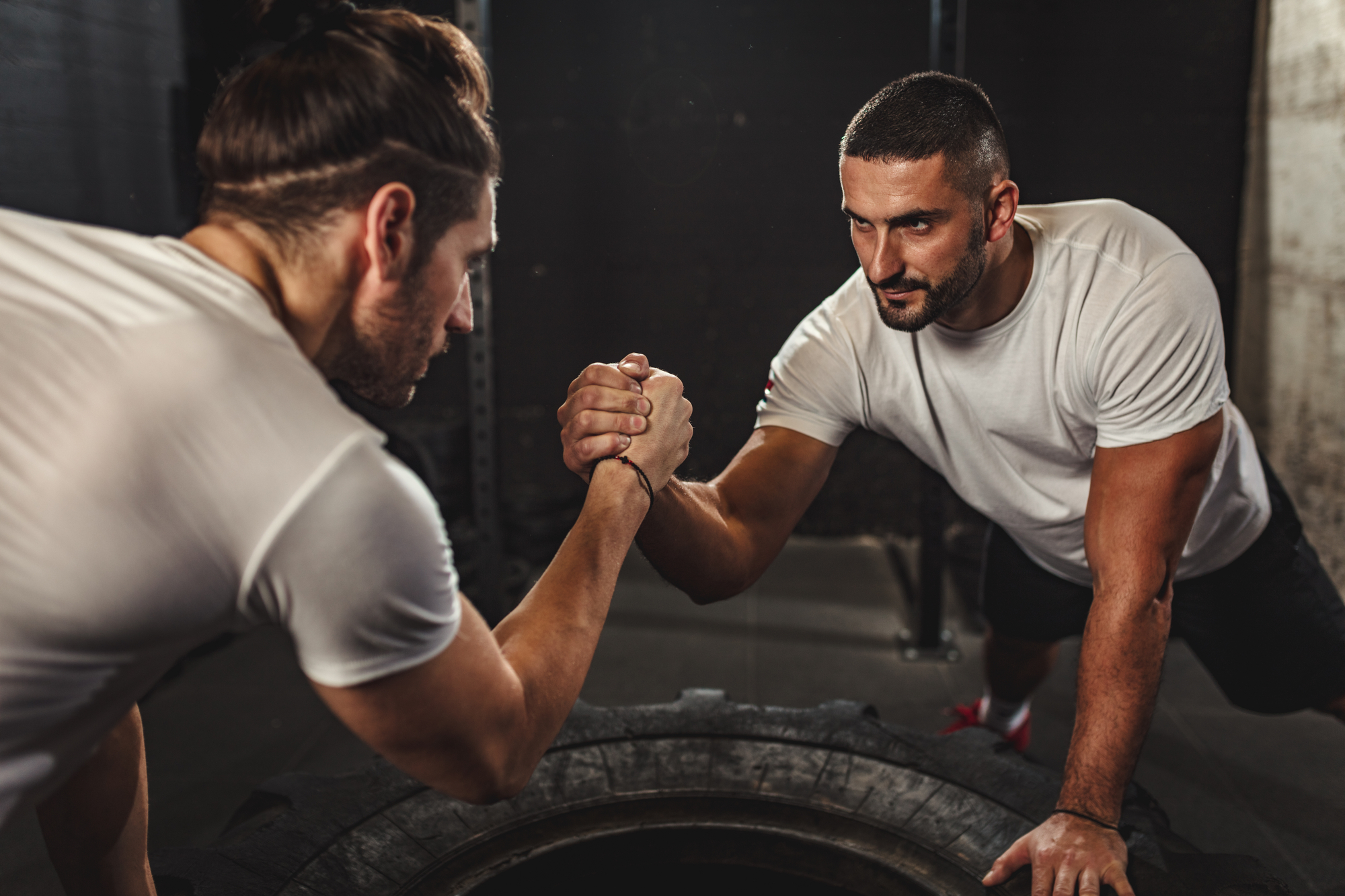 Two men, both wearing white t-shirts, engage in a competitive arm-wrestling match while leaning over a large tractor tire. They appear focused and determined. The background shows a dimly lit indoor gym setting with minimal equipment visible.