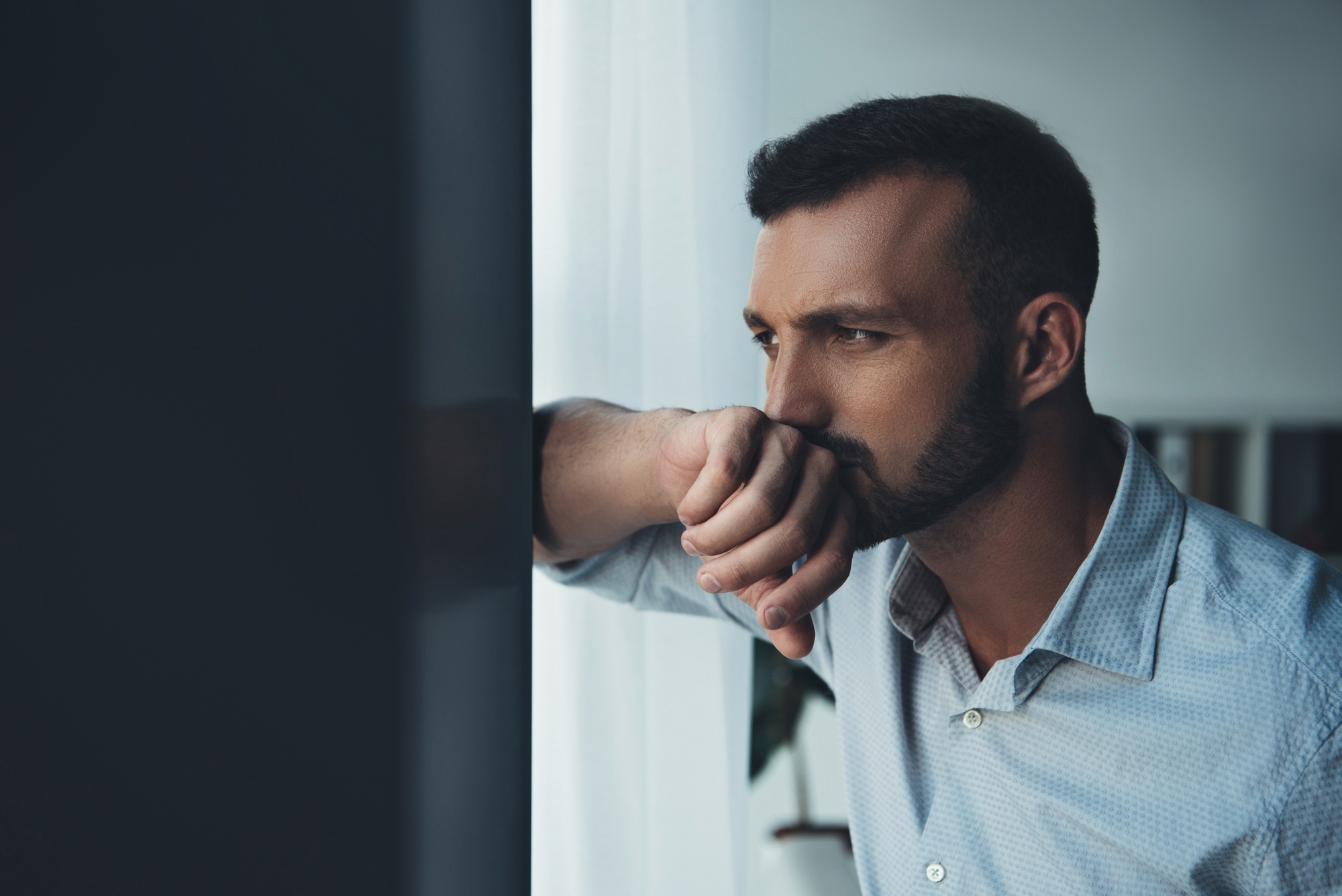 A man with short hair and a beard is standing indoors, looking out a window with a serious expression. He is resting his chin on his hand and wearing a light button-up shirt. The background is blurred, focusing on his contemplative posture.