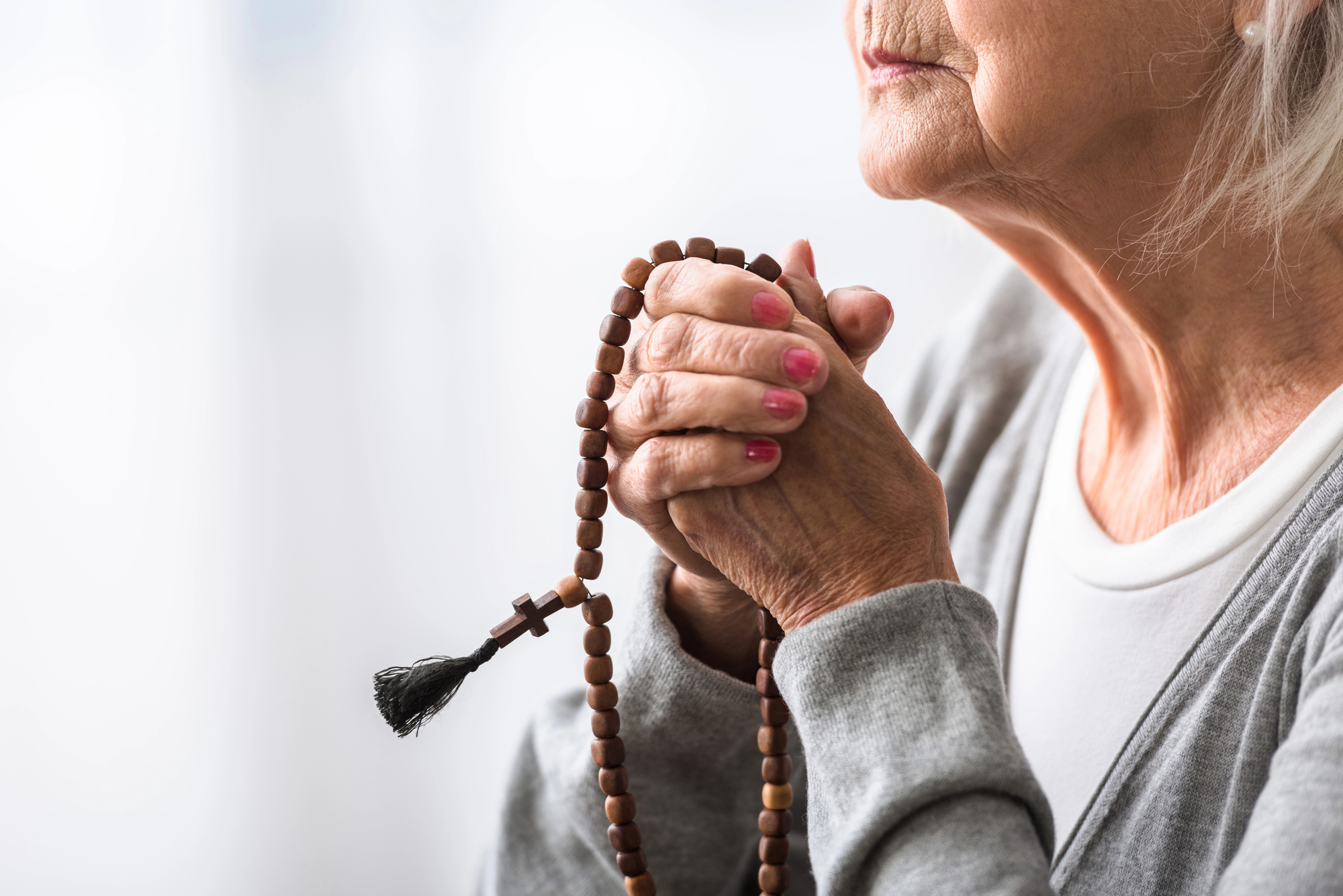 An elderly person holds a wooden rosary with both hands close to their face, appearing to be in prayer. The background is a softly lit, blurred white. The individual wears a grey sweater and a white shirt, with focus on their clasped hands and the rosary.
