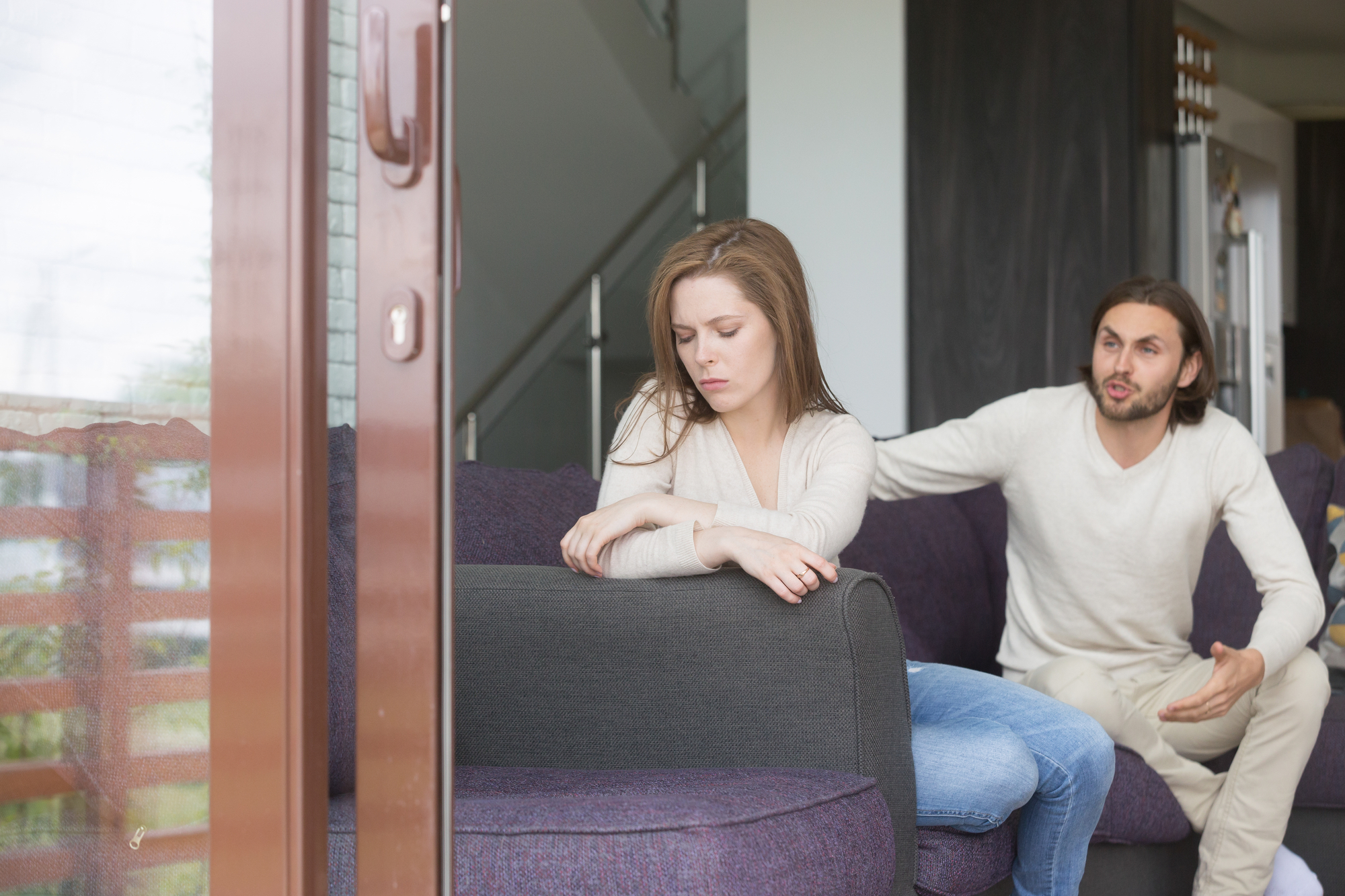 A woman sits on a grey couch looking down with a sad expression, while a man, also on the couch, gestures emphatically toward her with a frustrated look. They are in a modern living room with a glass door and a staircase in the background.