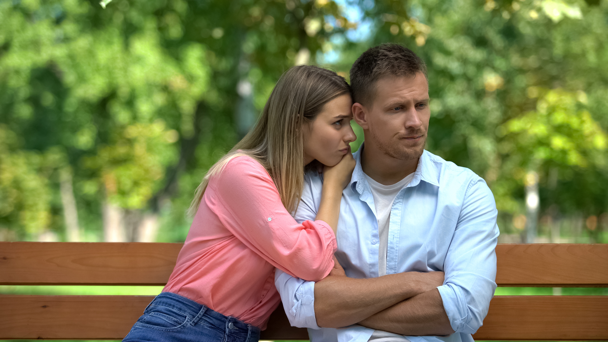 A woman with long hair in a pink shirt leans on a man in a light blue shirt who is sitting with crossed arms on a wooden bench. They both seem pensive and distant. The background displays lush green trees, likely in a park.