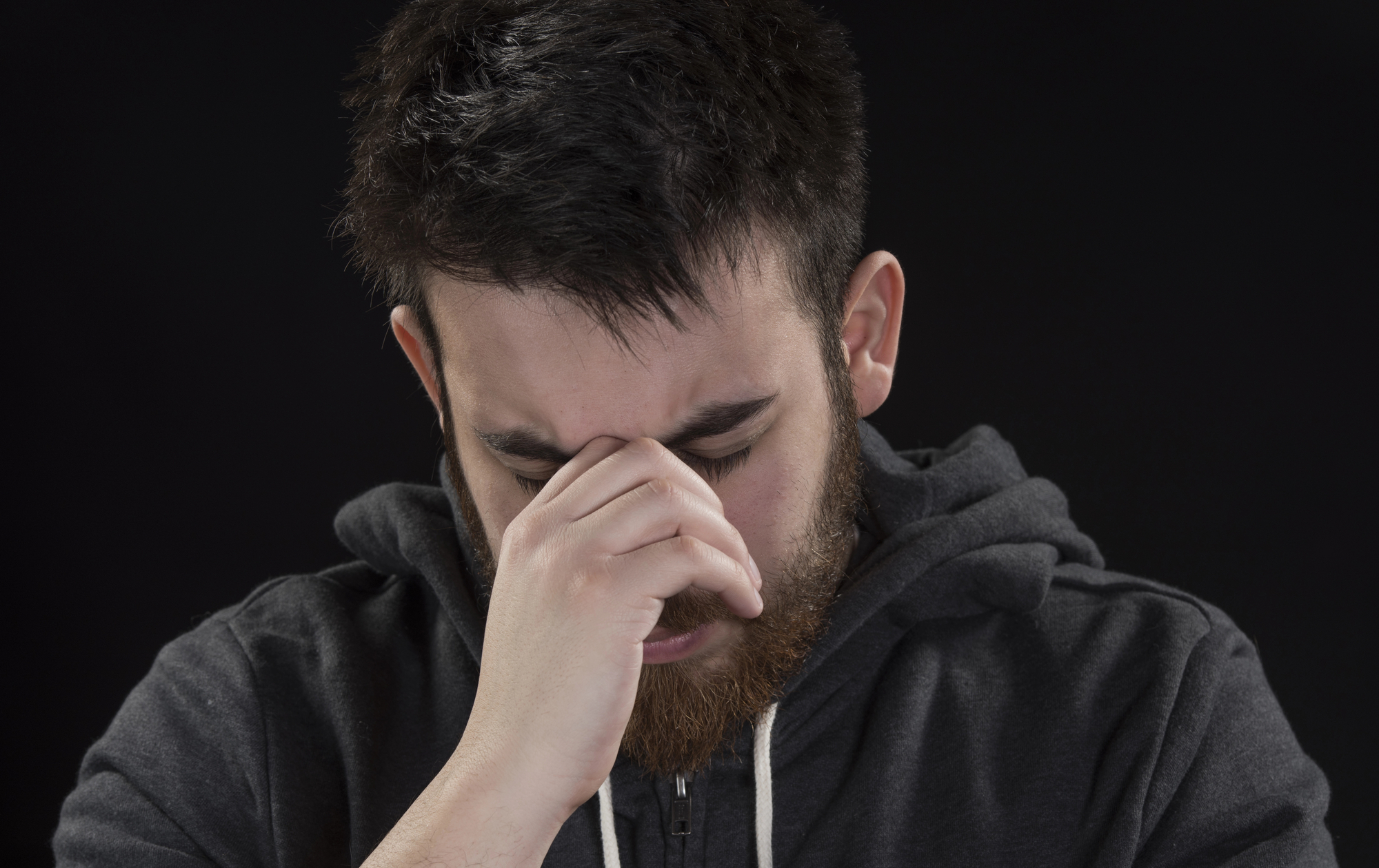 A man in a dark hoodie with a beard and short hair is shown against a dark background. He appears distressed, with his hand covering his nose and eyes closed, as if experiencing stress or frustration. The lighting is focused on his face, emphasizing his expression.
