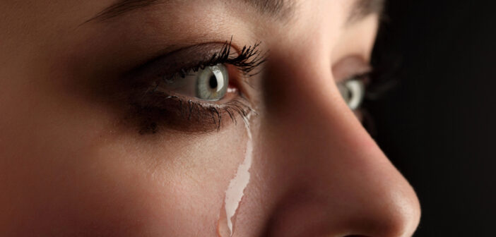 Close-up of a woman's face with a single tear rolling down her cheek. The image focuses on her expressive eyes with smeared eye makeup, reflecting an emotion of sadness or distress. The background is dark, highlighting her facial features.
