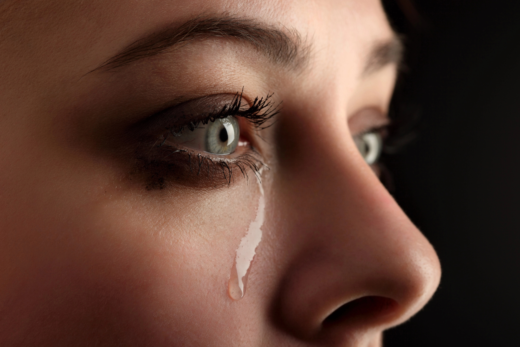 Close-up of a woman's face with a single tear rolling down her cheek. The image focuses on her expressive eyes with smeared eye makeup, reflecting an emotion of sadness or distress. The background is dark, highlighting her facial features.