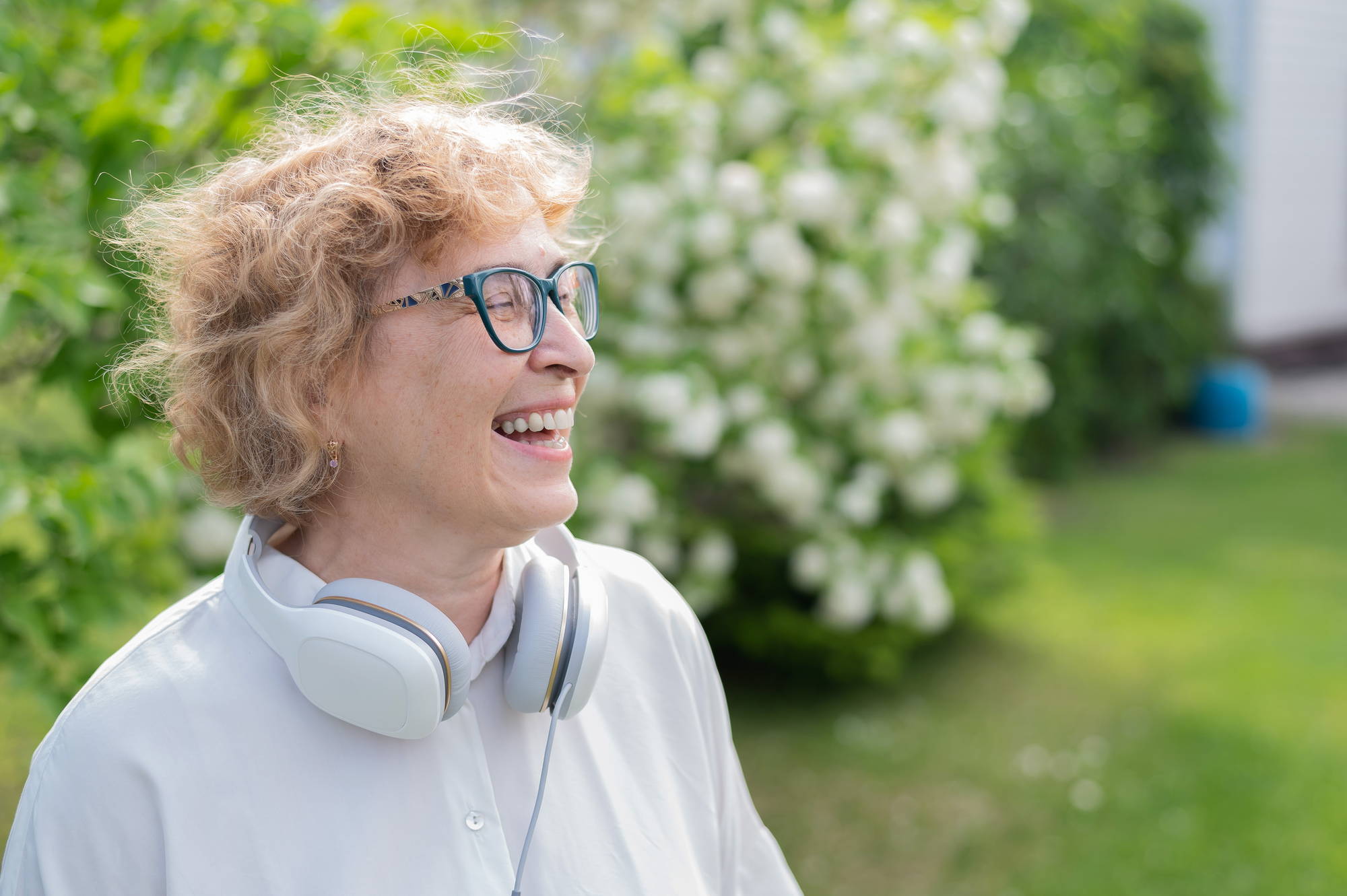 A smiling person wearing glasses and a white shirt stands outside. They have curly hair and are wearing headphones around their neck. The background is lush and green, with blooming white flowers.