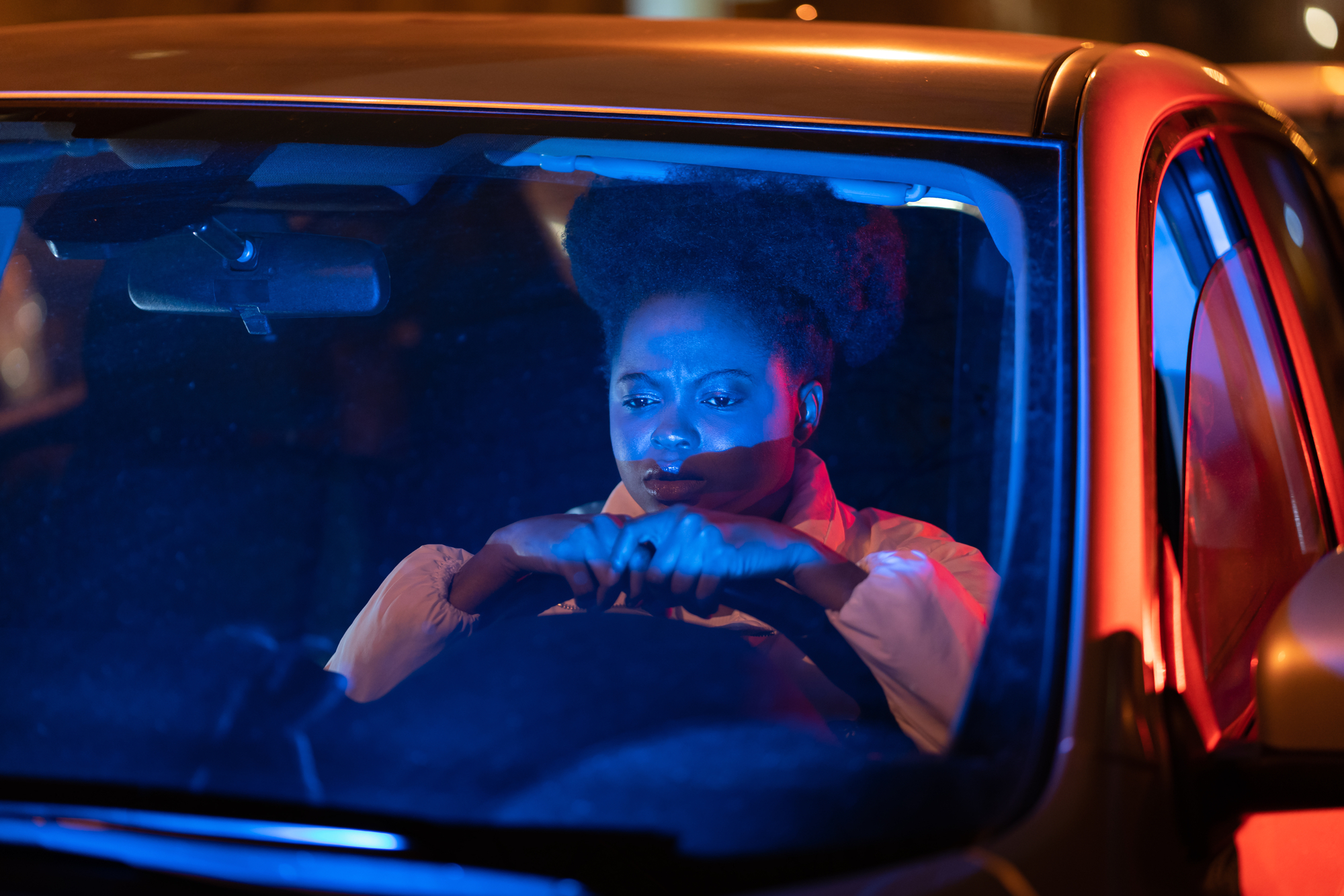 A person with an afro hairstyle is seen through the windshield of a car at night. They appear concerned, gripping the steering wheel tightly. The scene is illuminated by blue and red lights, suggesting the presence of emergency or police vehicles nearby.