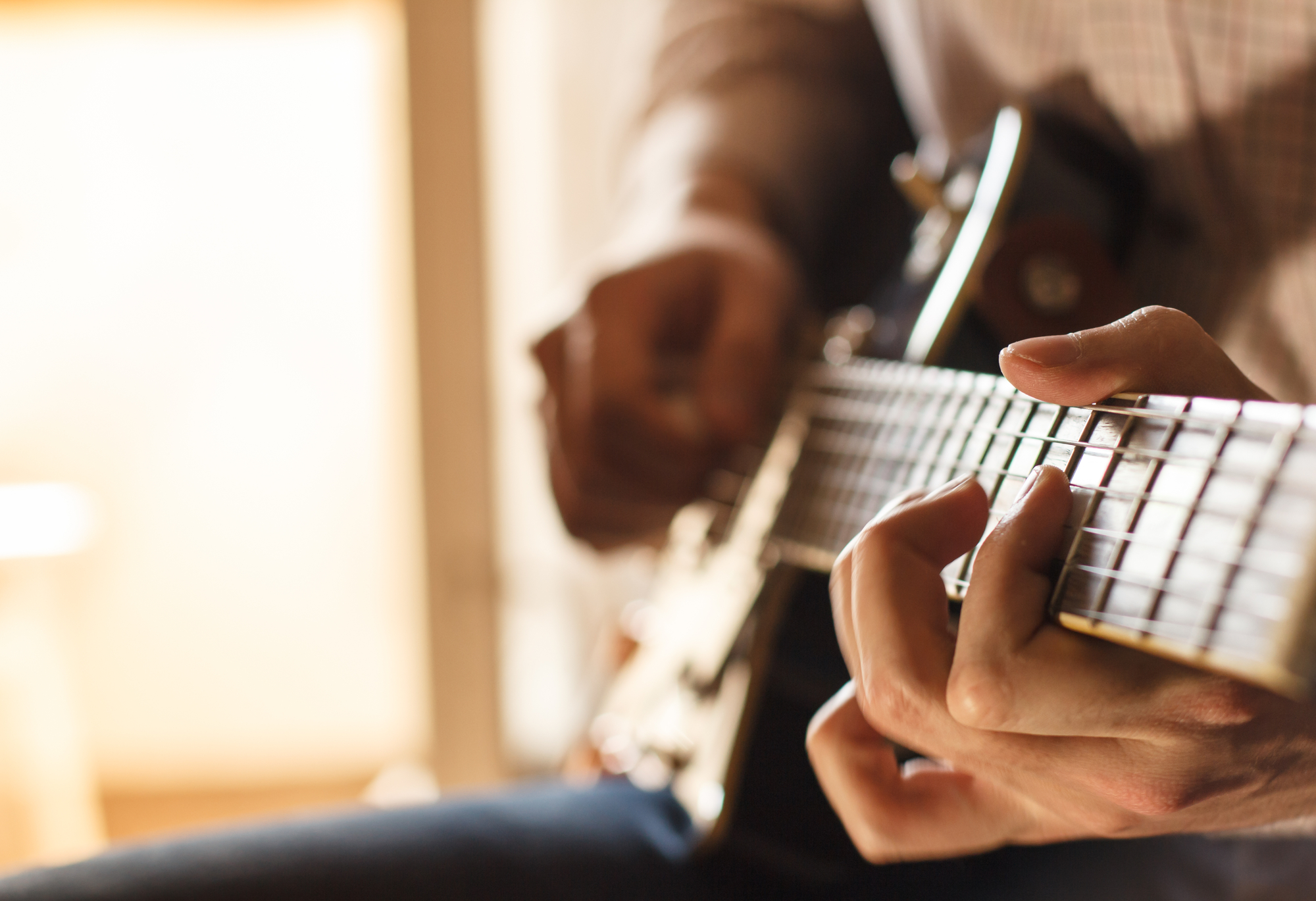 Close-up of a person’s hands playing an electric guitar. The left hand is pressing down on the strings on the fretboard while the right hand is positioned near the body of the guitar, likely strumming or picking the strings. The background is softly blurred.