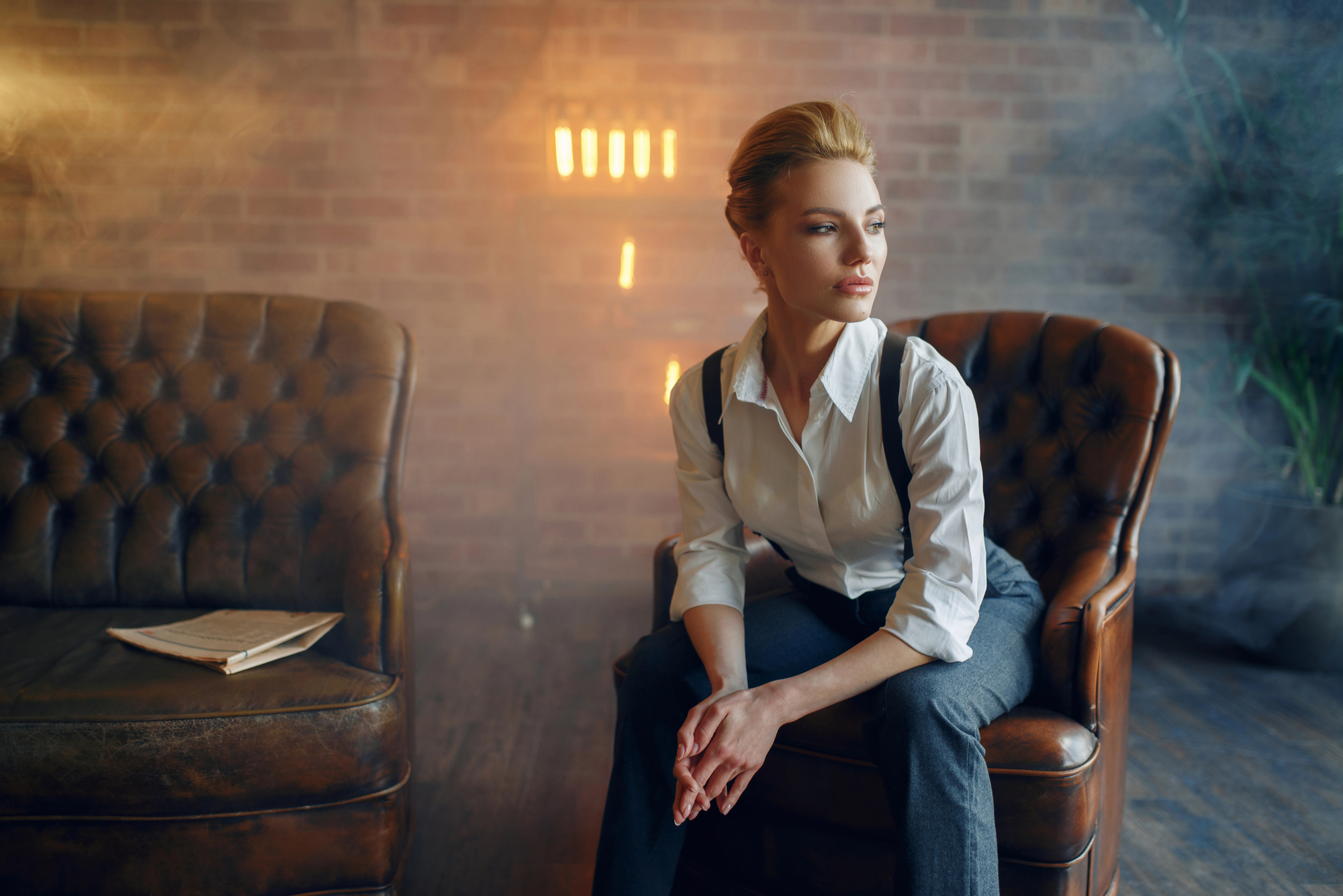 A woman with blond hair, dressed in a white shirt and dark pants, sits pensively on a leather armchair. Another empty leather armchair is to her left, with a document on it. The background has a brick wall and hazy lighting, giving a moody atmosphere.