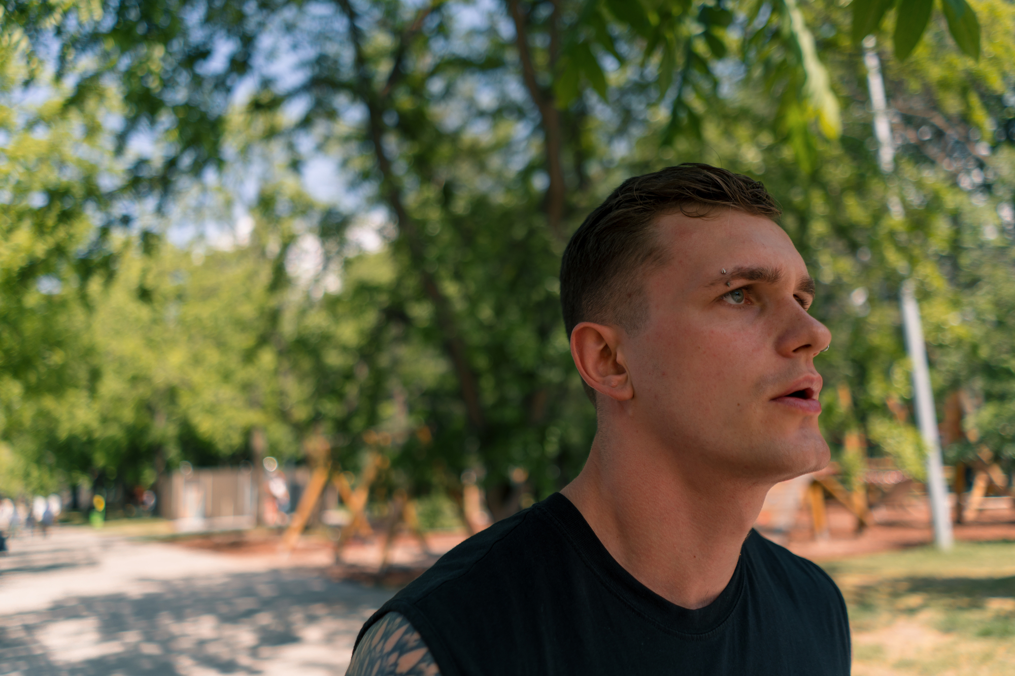 A young man with short hair and a tattoo on his left arm is standing outdoors, looking to the side with a thoughtful expression. He is wearing a black sleeveless shirt. The background is a sunlit park with trees, grass, and a dirt path.