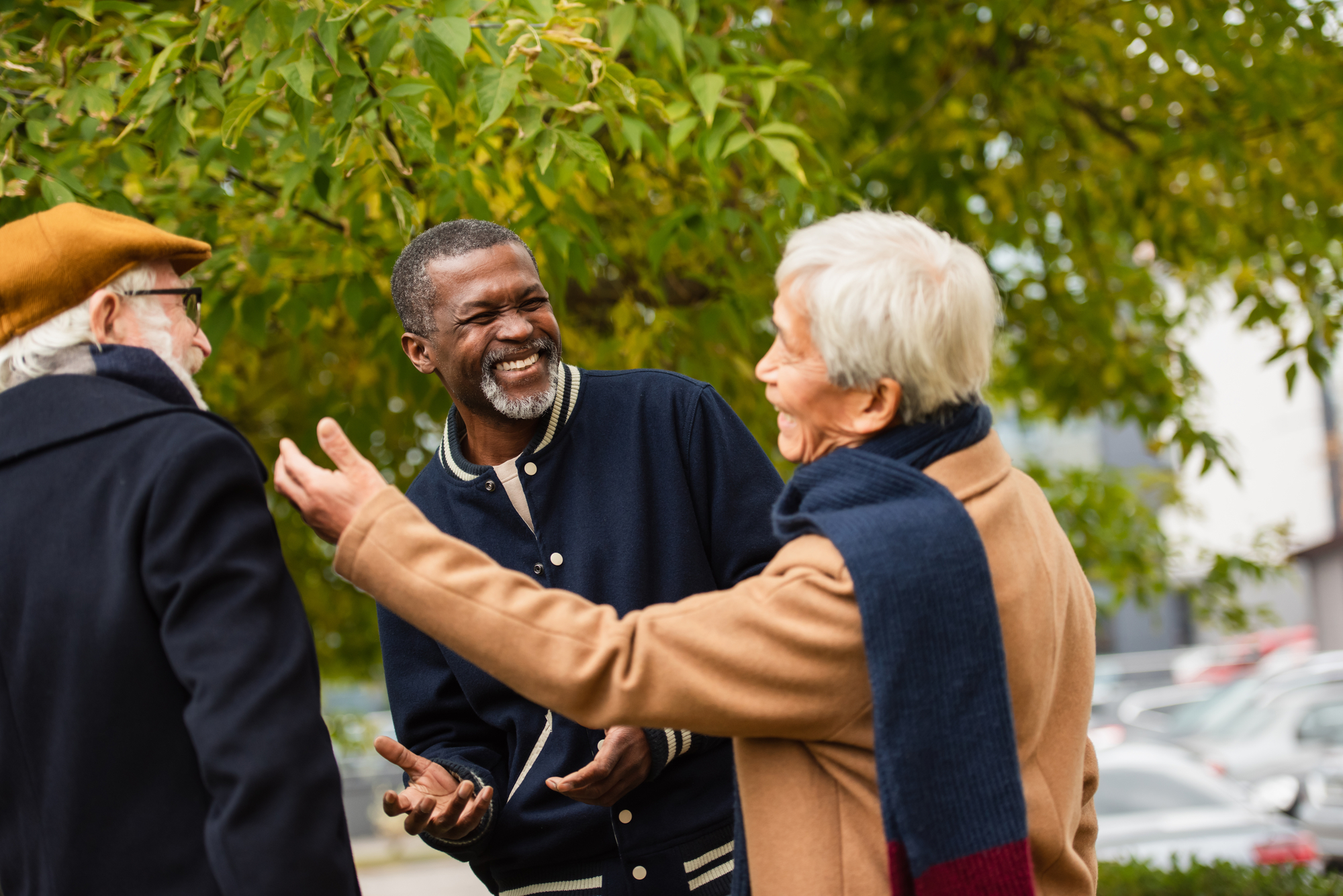 Three elderly people are outdoors, warmly dressed in coats and scarves. They are standing under a tree with green leaves and appear to be having a lively conversation. The person in the middle is laughing, creating a joyful atmosphere among the group.