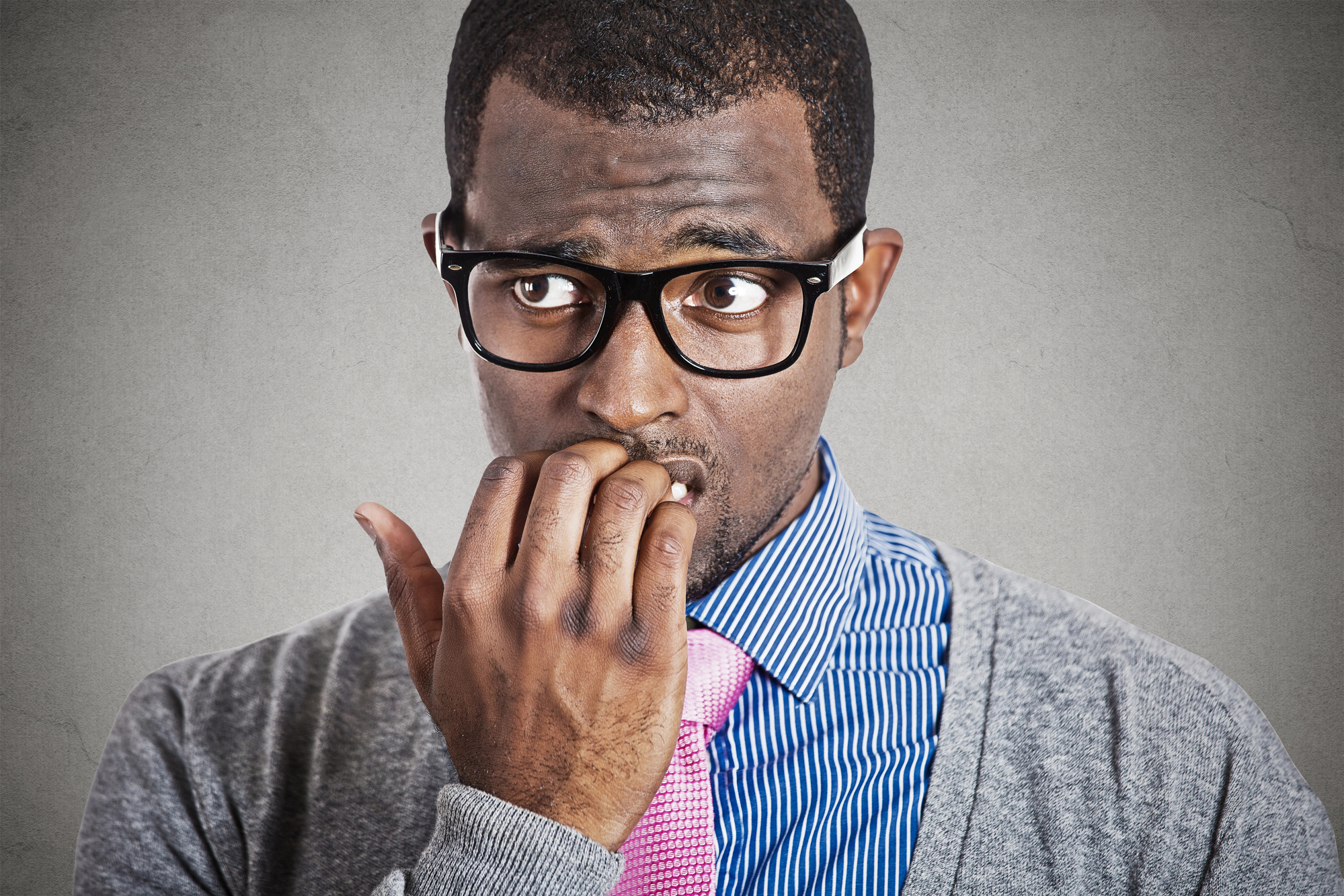 A man wearing glasses, a striped blue shirt, a pink tie, and a gray sweater is looking to his right with a concerned expression, biting his nails, against a gray background.