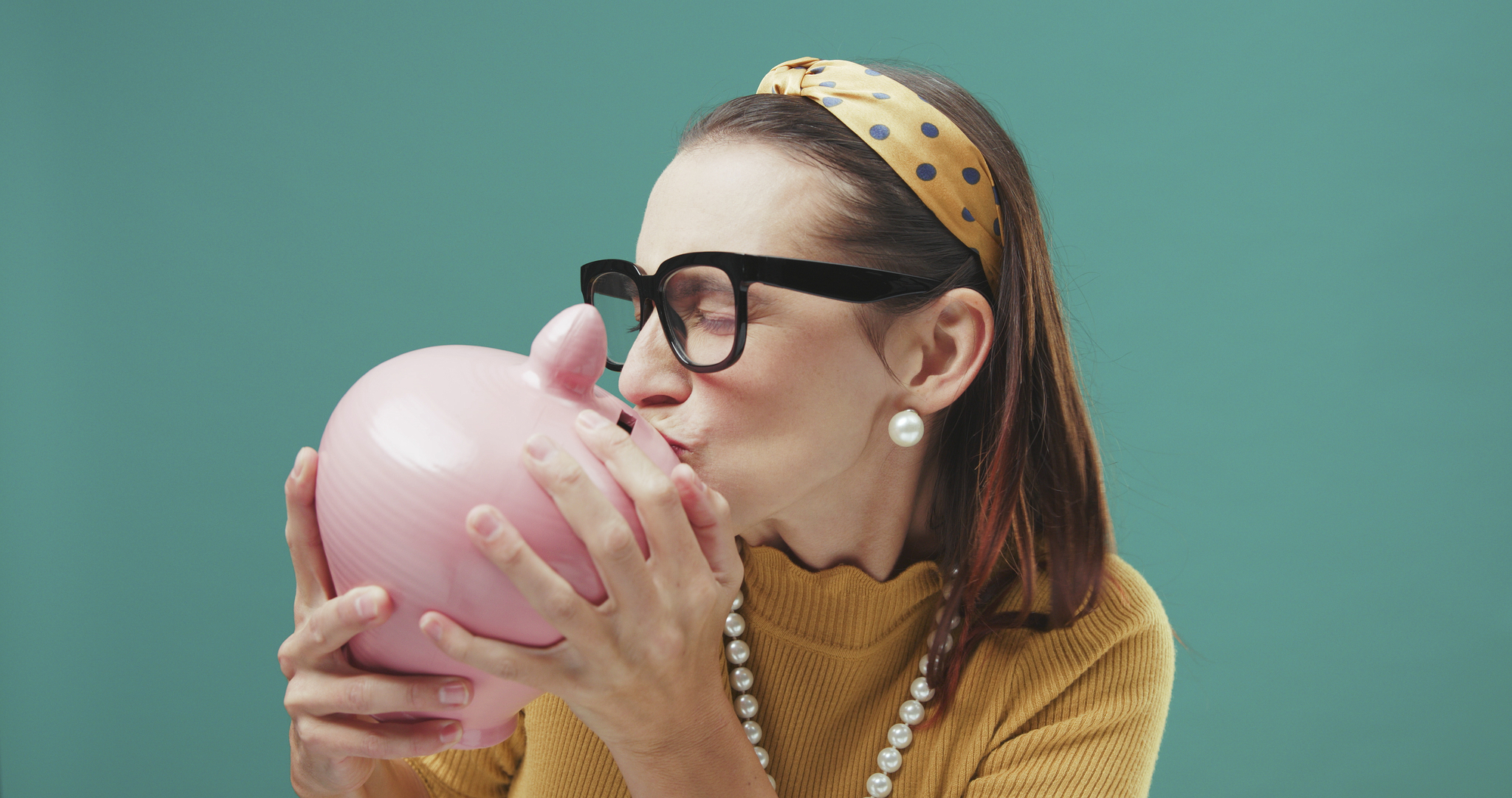 A woman wearing a yellow headband with polka dots, large black glasses, and a mustard-colored sweater is kissing a pink piggy bank. She is also wearing pearl earrings and a matching necklace. The background is a solid teal color.