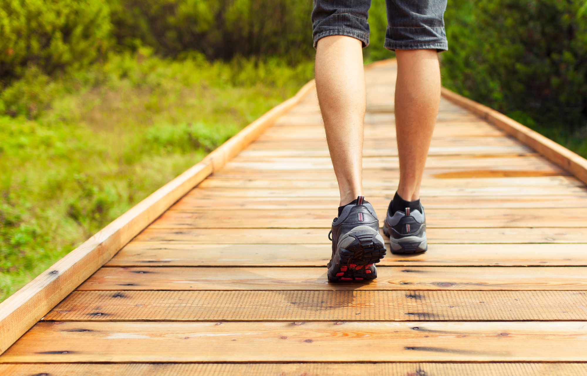 A person's legs in athletic shoes walking on a wooden pathway in an outdoor setting with greenery on both sides. The person is wearing dark shorts and appears to be enjoying a hike or walk in nature.