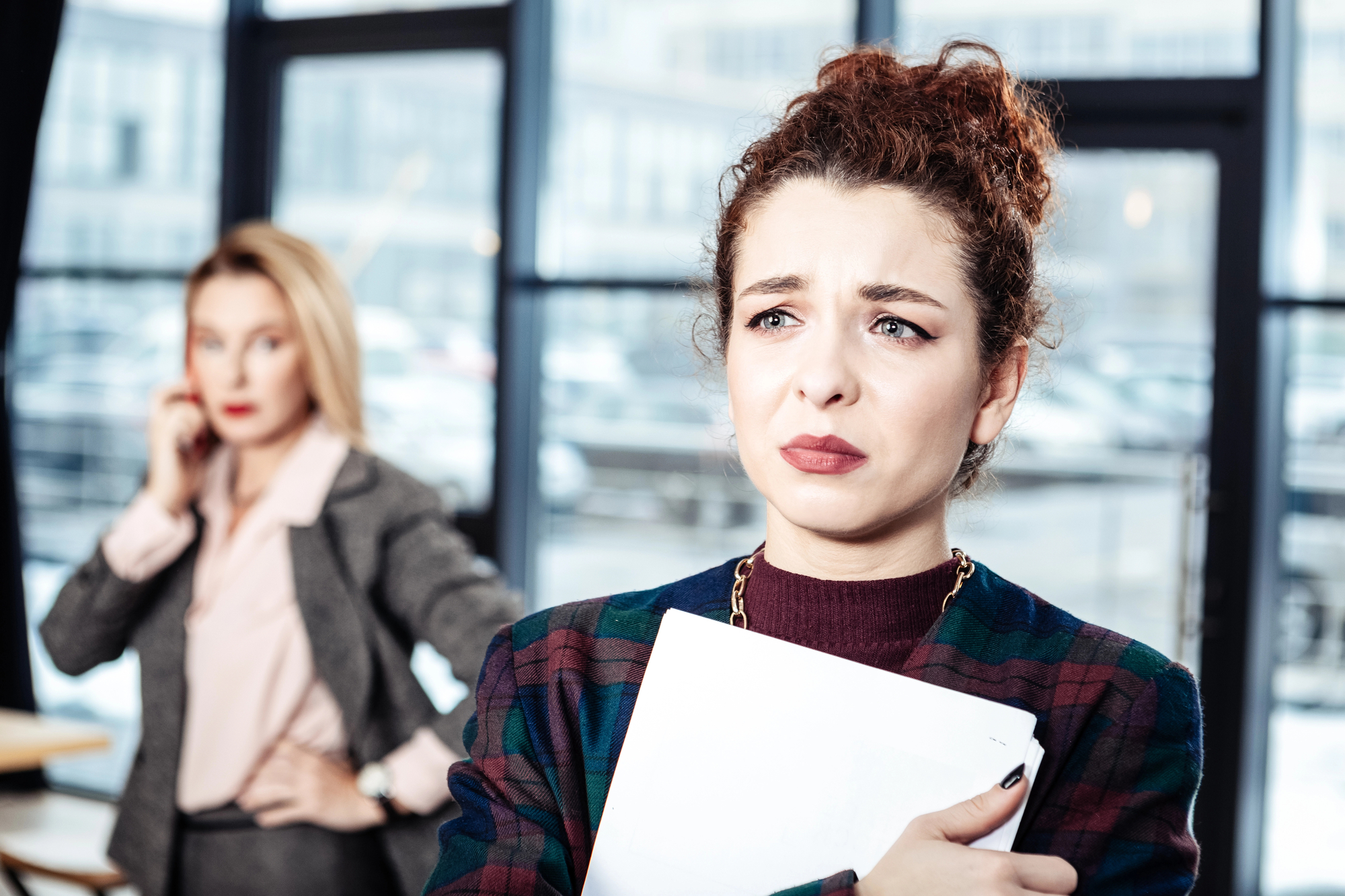 A woman with curly hair and a concerned expression holds a clipboard while standing in the foreground. In the background, another woman with blonde hair and a serious expression talks on the phone. They are in a modern office space with large windows.