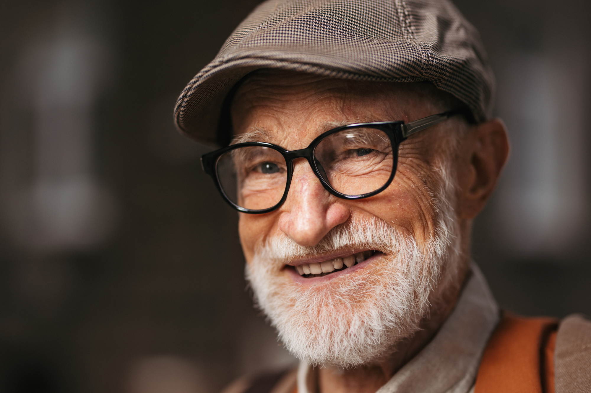 An older man with a white beard and glasses is smiling warmly. He is wearing a flat cap and a brown jacket. The background is blurred, focusing attention on his joyful expression.