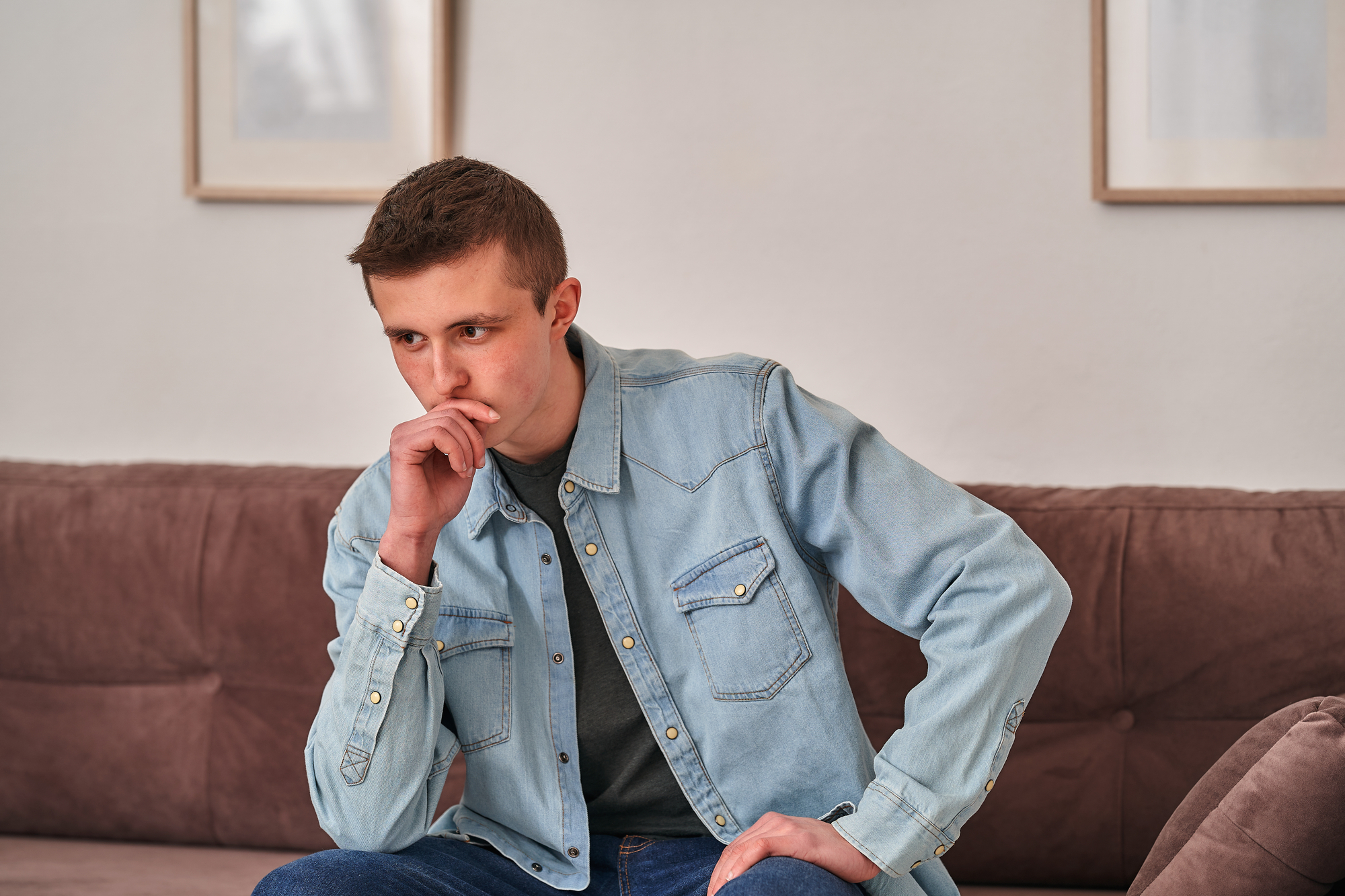 A young man sits on a brown couch in a relaxed home setting, leaning forward with his elbow on his knee and his hand covering his mouth in a thoughtful pose. He is wearing a light blue denim shirt over a gray t-shirt. The background features two framed pictures.