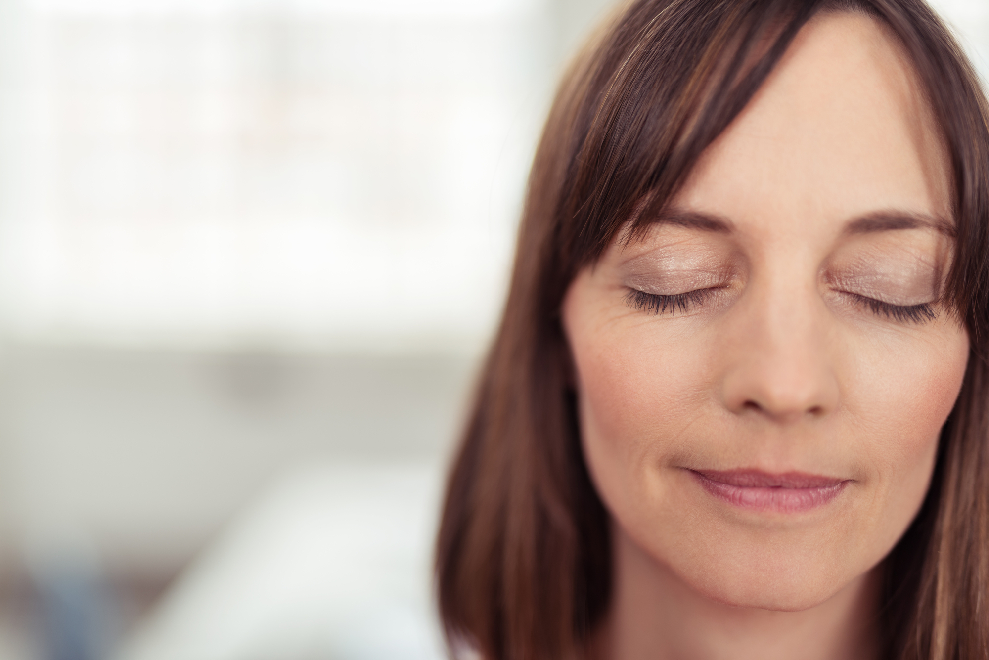 A woman with shoulder-length brown hair and bangs is shown in a close-up with her eyes closed and a serene expression on her face. The background is out of focus, suggesting she might be indoors.