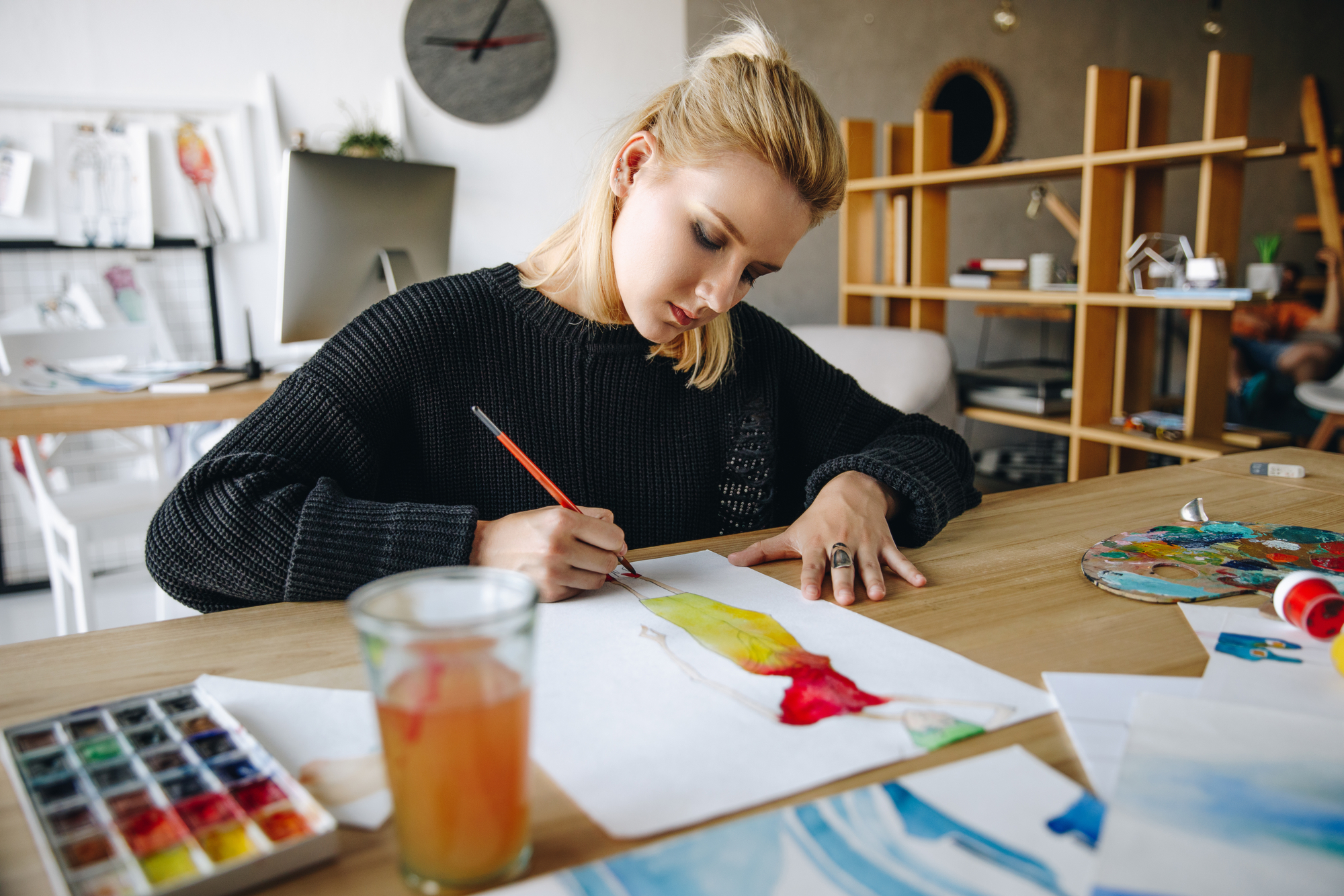 A person with blonde hair, dressed in a black sweater, is seated at a desk, focused on painting. Various art supplies surround them, including watercolor paints, a glass of water, and other paper works. Shelving and a clock are visible in the background.