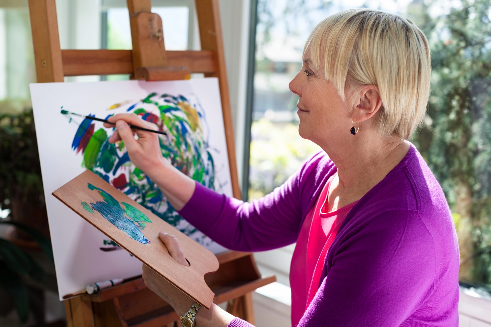 An older woman with short blonde hair, wearing a bright pink cardigan, is painting on a canvas in a well-lit room. She holds a palette in her left hand and a brush in her right, focusing intently on her colorful abstract artwork.
