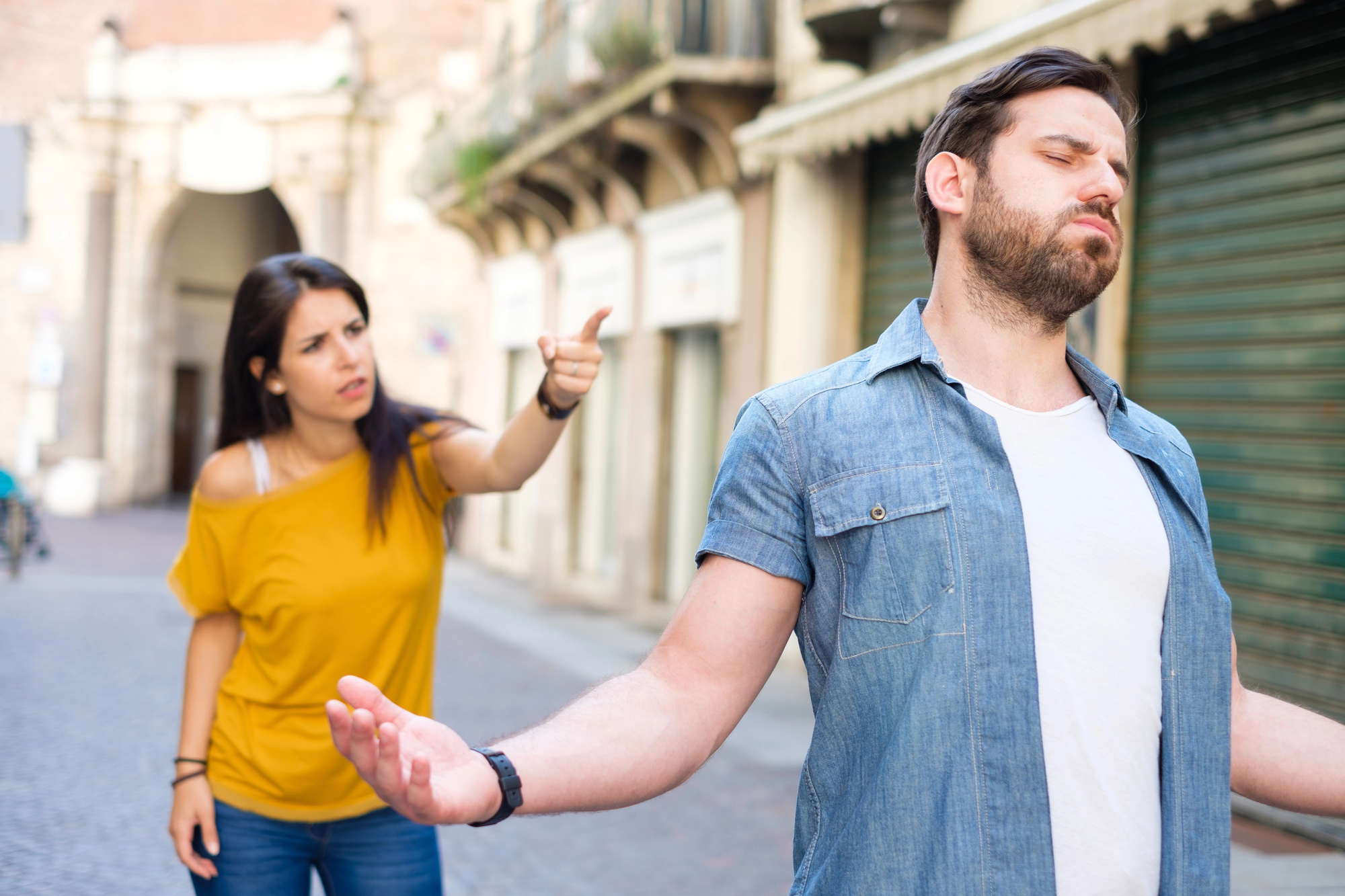 A woman in a yellow top is pointing and shouting at a bearded man in a denim shirt. The man has his back turned to her, his eyes closed, and his arms slightly raised, looking frustrated. They are standing on a cobblestone street with buildings in the background.