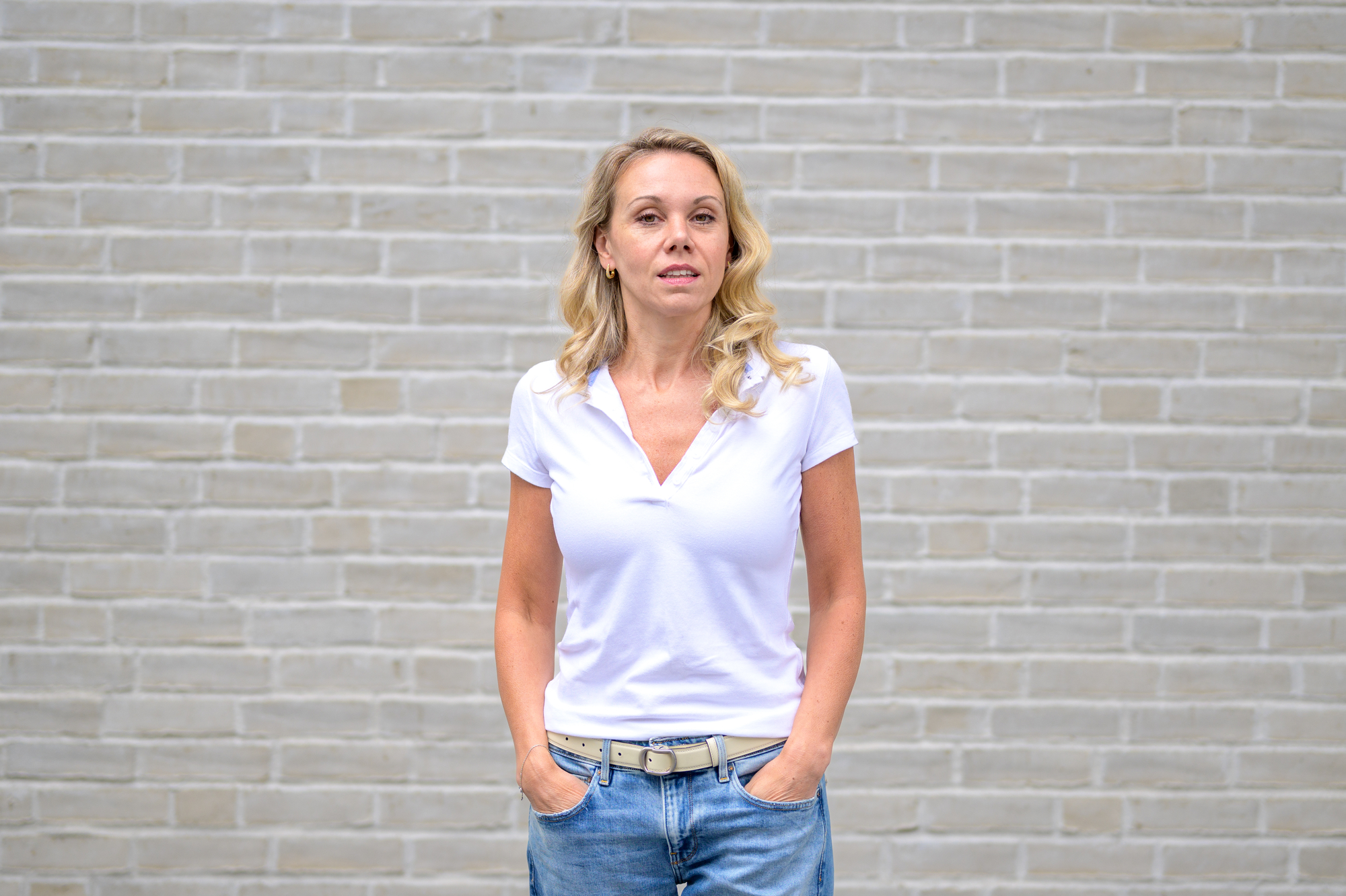A woman with light-colored hair stands in front of a light gray brick wall. She is wearing a white short-sleeve polo shirt, blue jeans, and a light-colored belt. She has her hands in her pockets and is looking directly at the camera.
