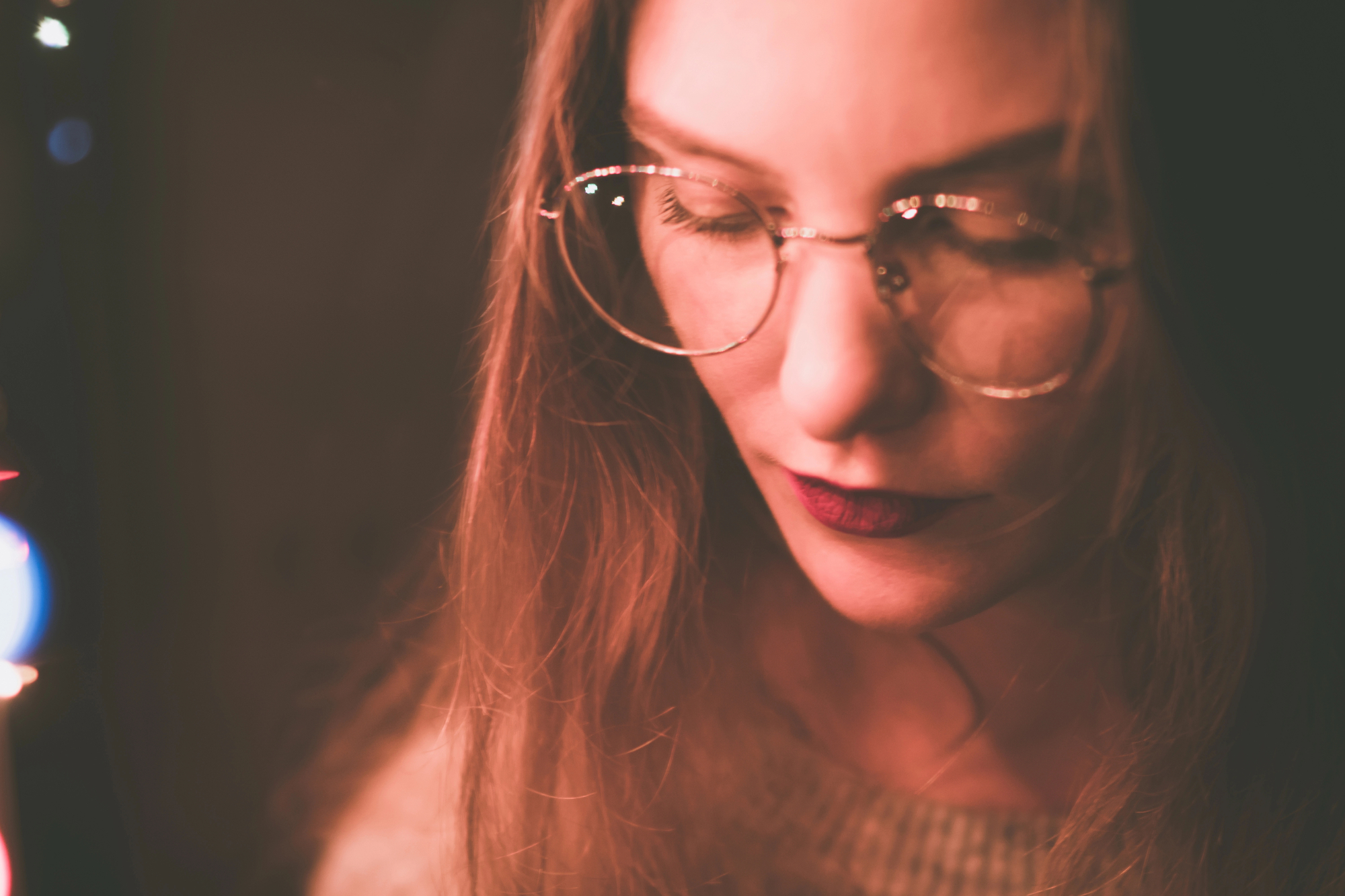 Close-up of a woman with long hair, wearing round glasses. She is looking down, with a serious or thoughtful expression. The lighting is soft and warm, casting a gentle glow on her face. The background is dark and blurred.