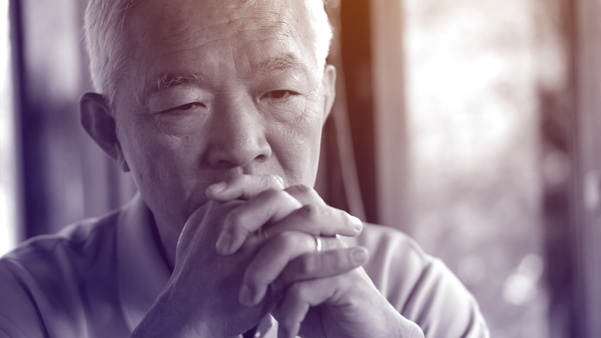A pensive elderly man with white hair rests his chin on his folded hands, gazing thoughtfully into the distance. The background is softly blurred with natural light coming through a window. The image has a gentle, sepia tone.