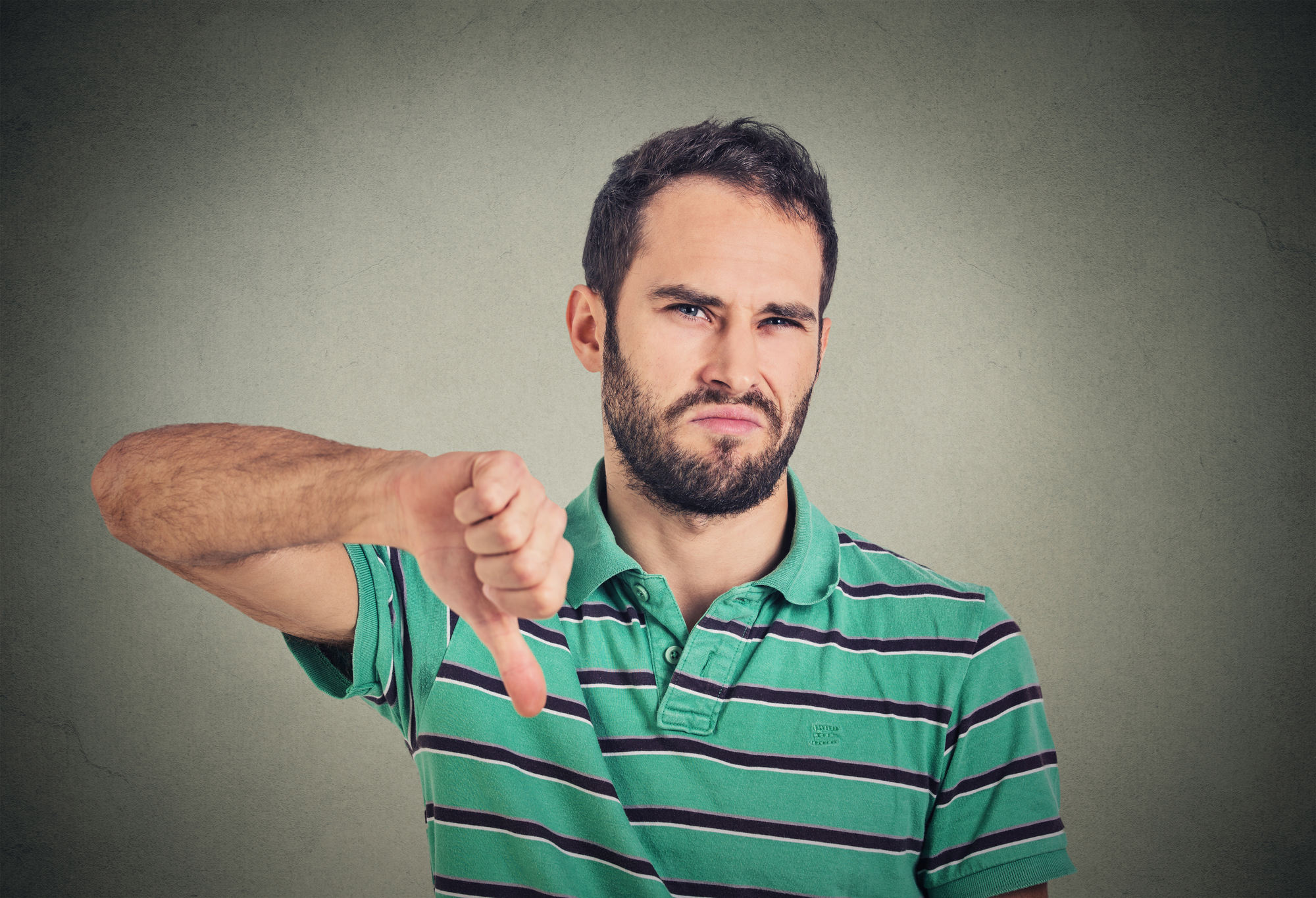 A man with a beard wearing a green and white striped polo shirt gives a thumbs-down gesture with his right hand, while his face displays a displeased expression. The background is a plain gray wall.