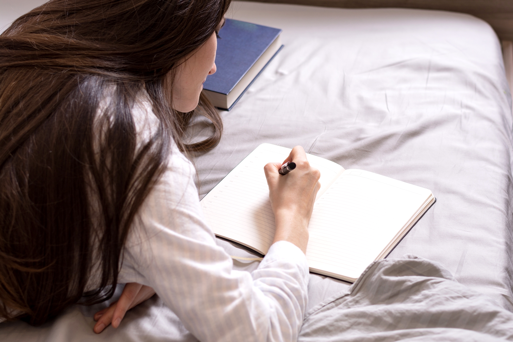 A person with long brown hair, dressed in a light-colored shirt, is lying on a bed while writing in a notebook with a black pen. The bed has grayish bedding and an open book lies next to them. The scene appears calm and focused.