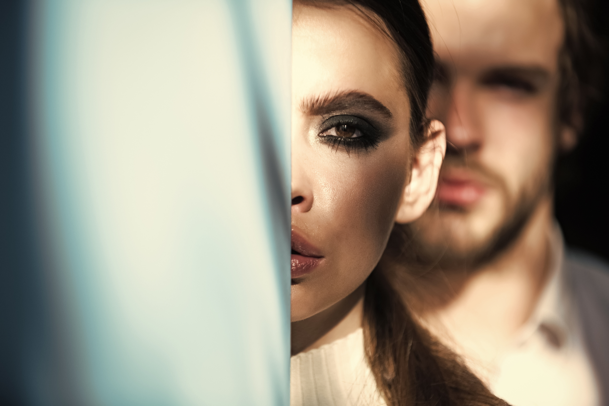 Close-up of a woman partially obscured by a blue object, leaving only one heavily made-up eye visible. Behind her, a man with a beard is blurred and partially visible. The lighting casts a dramatic shadow across both faces.