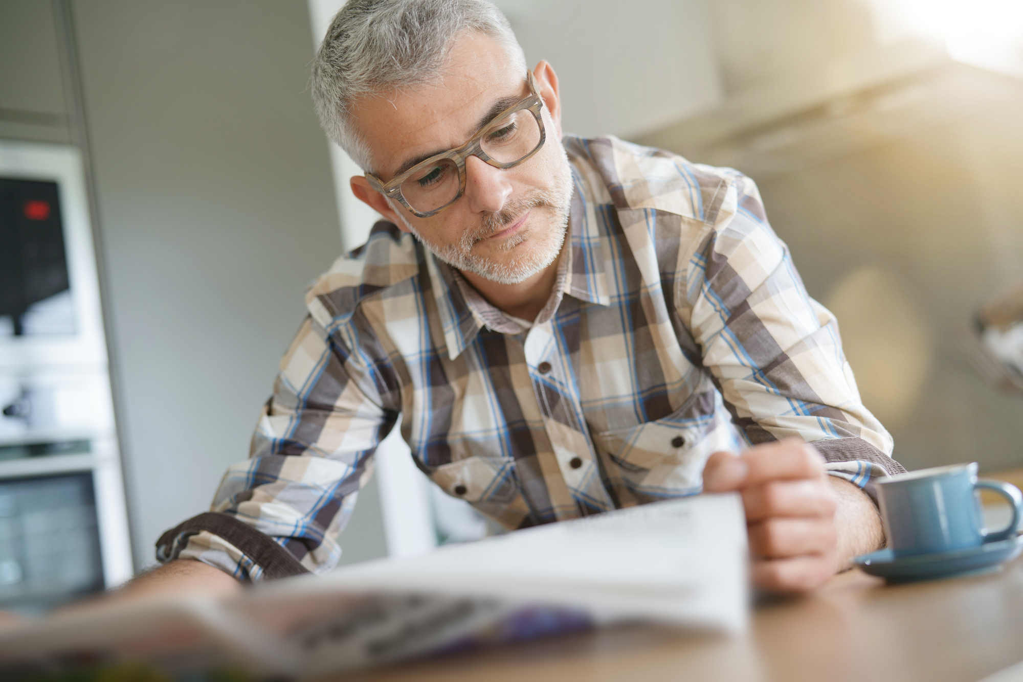A middle-aged man with gray hair and glasses reads a newspaper at a table. He is wearing a checkered shirt and is focused on the content. A blue coffee cup is placed on the table next to him, and sunlight is streaming into the room.