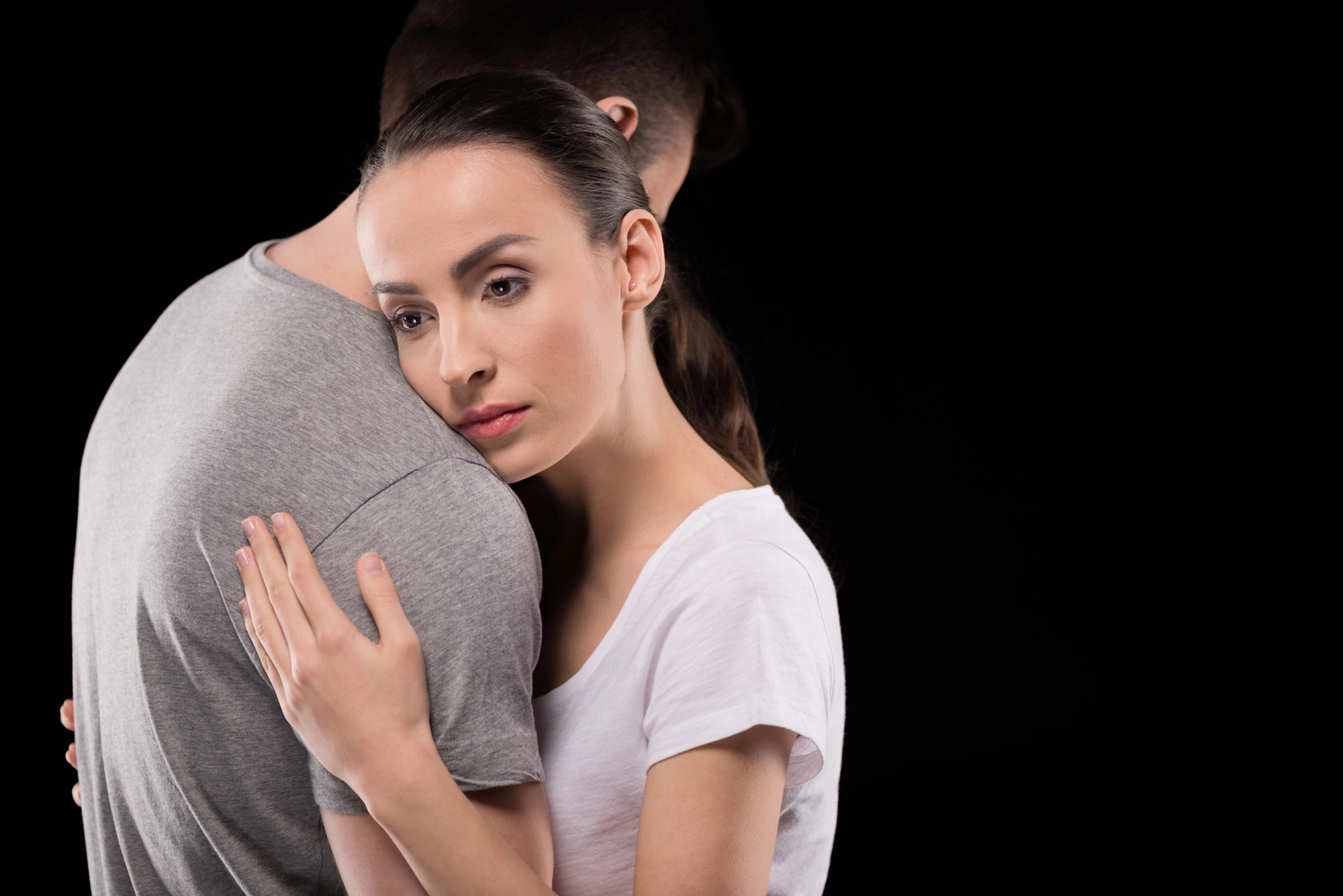 A woman in a white shirt embraces a man in a gray t-shirt from behind, resting her head on his shoulder. They are against a black background, and the woman has a contemplative expression. The man's face is not visible.