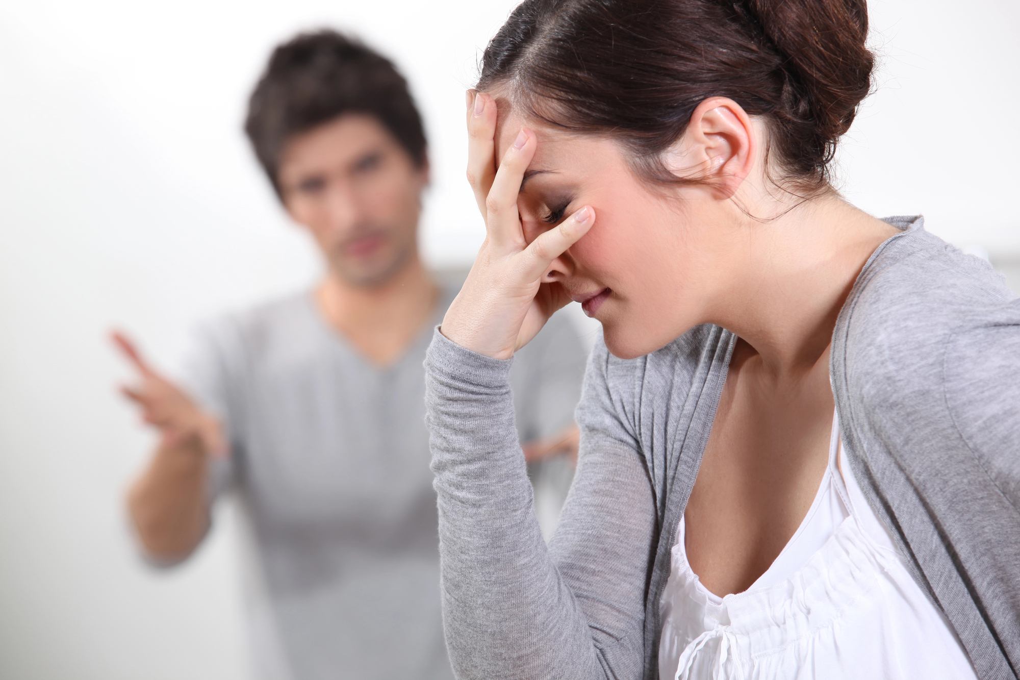 A woman in focus stands in the foreground with her hand covering her face, appearing distressed. In the blurred background, a man is visible with his arms extended, seemingly in a heated conversation. Both are wearing casual clothing.