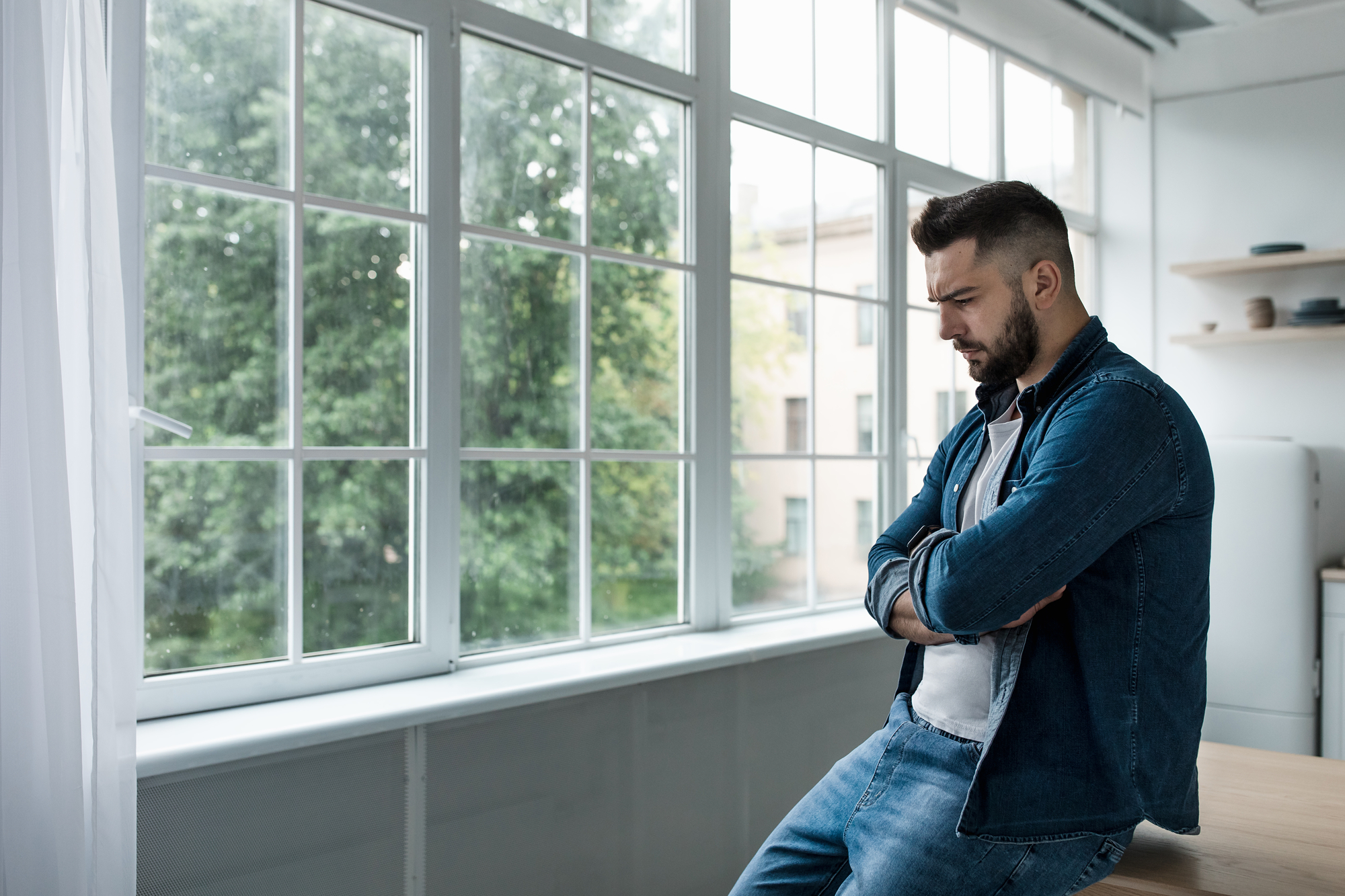 A man with a beard and short hair stands pensively by a large window with greenery visible outside. He is wearing a denim jacket over a white shirt and jeans, with his arms crossed and eyes looking down. The interior is bright with natural light from the window.