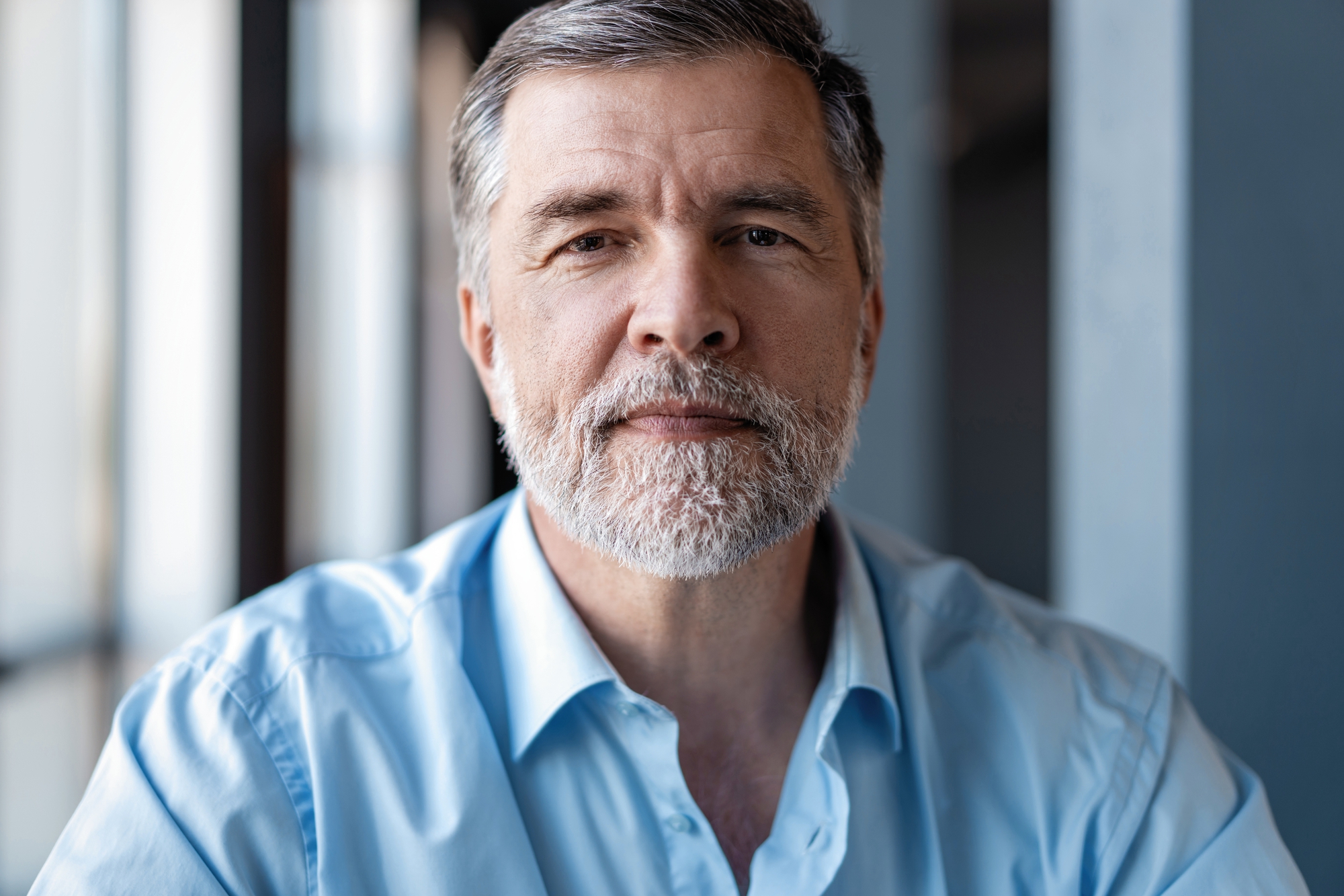 A middle-aged man with short, gray hair and a neatly trimmed beard is looking directly at the camera. He is wearing a light blue button-up shirt and appears to be indoors, possibly in an office or home setting, with soft natural light illuminating his face.