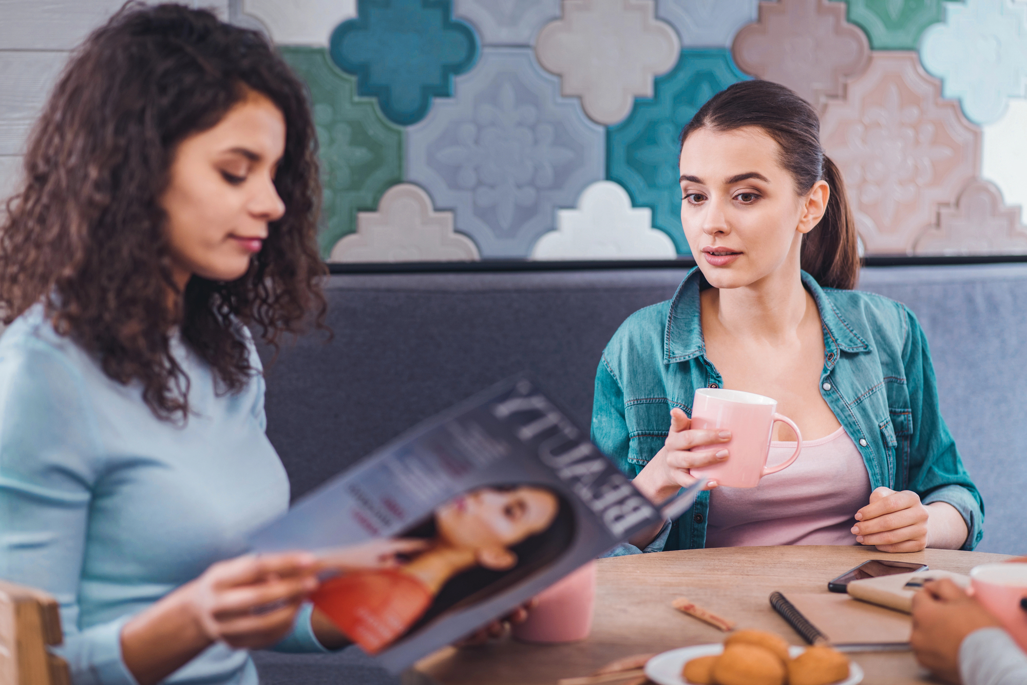 Two women sit at a table; one reads a fashion magazine while the other holds a pink mug and looks at her. They are in a cozy setting with colorful patterned wall tiles in the background. On the table are cookies and a smartphone.