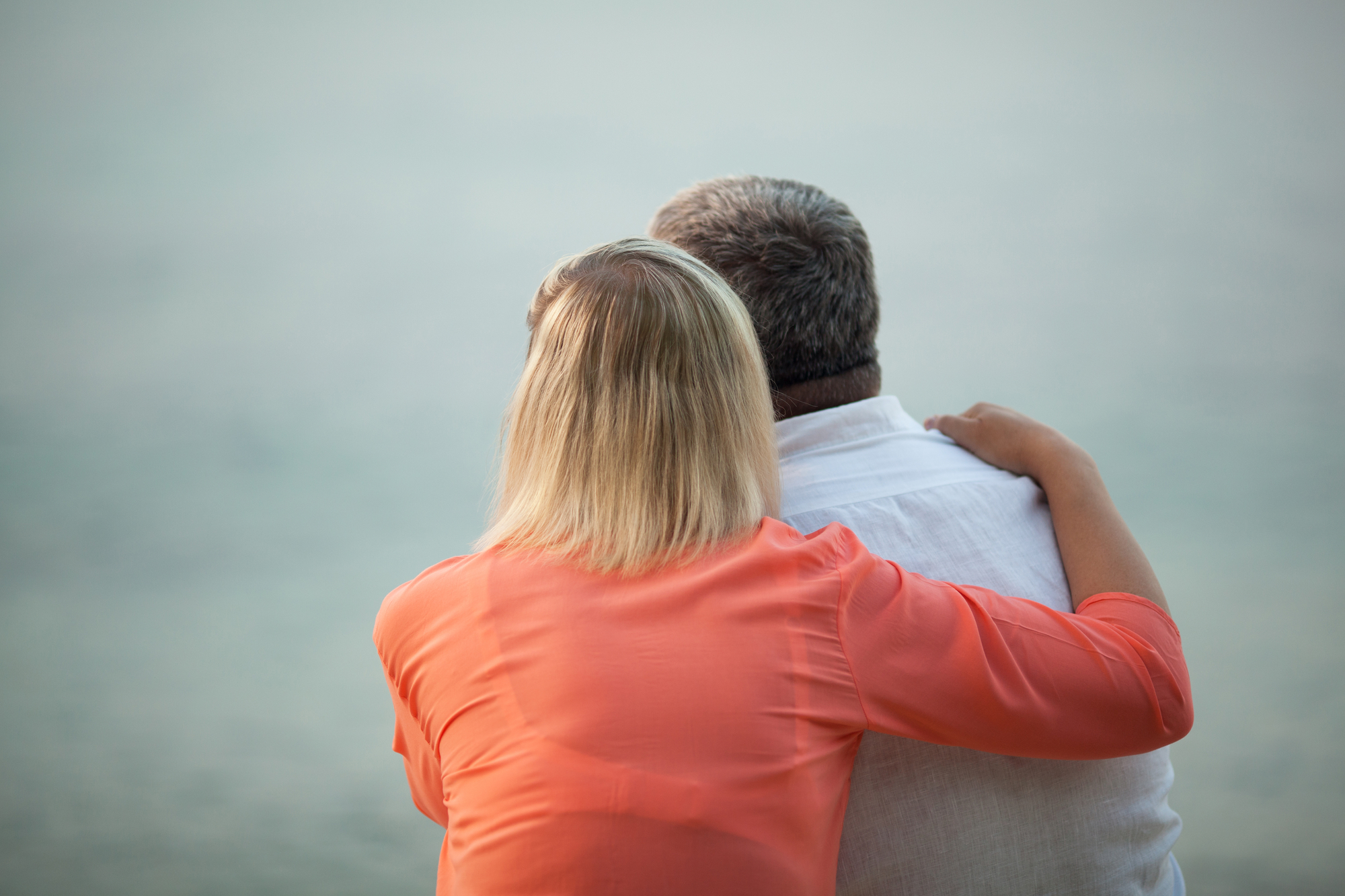 Two people sit closely together, viewed from behind, gazing at a calm body of water. The person on the left has shoulder-length blonde hair and wears an orange shirt, while the person on the right has short gray hair and wears a white shirt.