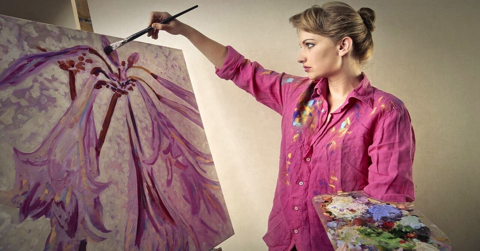 A woman wearing a pink shirt is painting on a large canvas. She is holding a palette in one hand and a brush in the other, adding details to her abstract artwork with purple and pink tones. The background is neutral, putting focus on the artist and her painting.