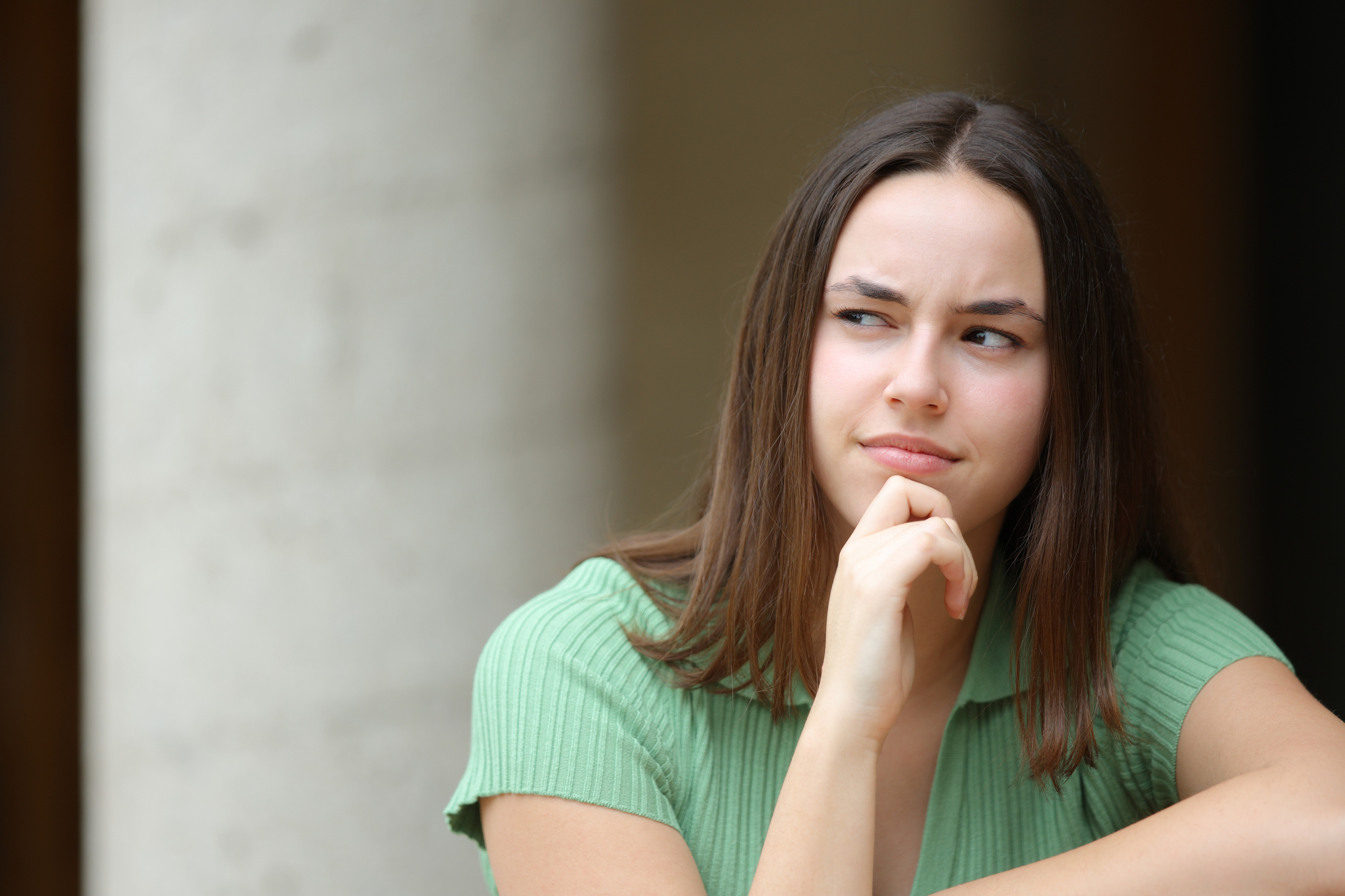 A young woman with long brown hair is resting her chin on her hand, appearing contemplative or uncertain. She is wearing a light green short-sleeved top and gazing slightly to the side. The background is out of focus.