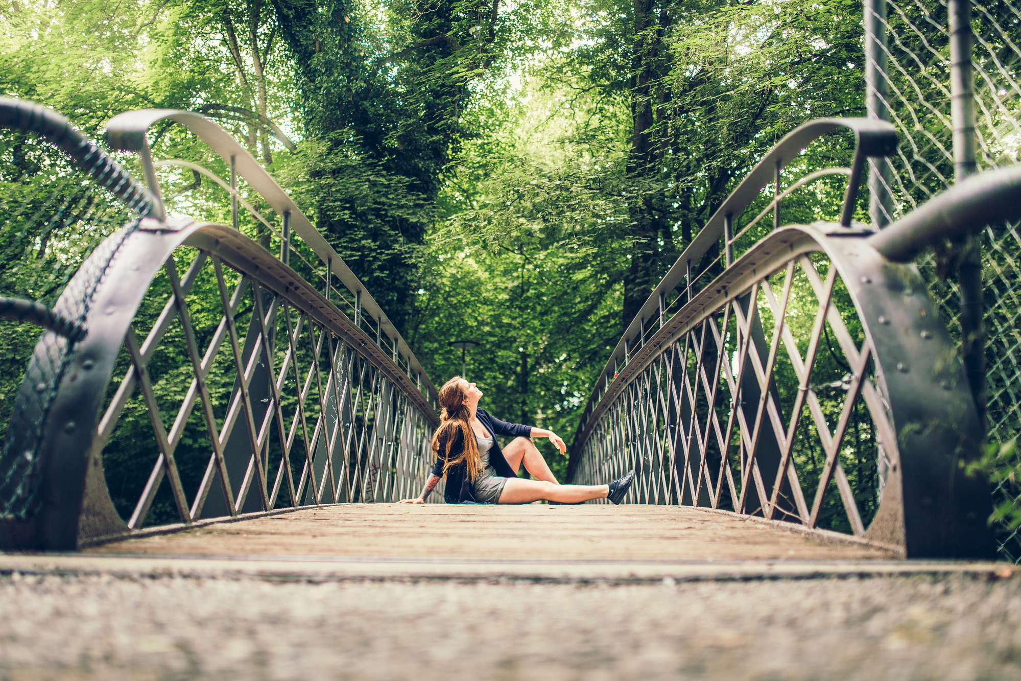 A young person with long hair, dressed casually, sits on the wooden planks of an arched metal bridge in a lush forest. They are looking at the greenery above, surrounded by tall trees and dense foliage. The perspective of the photo makes the bridge appear expansive.