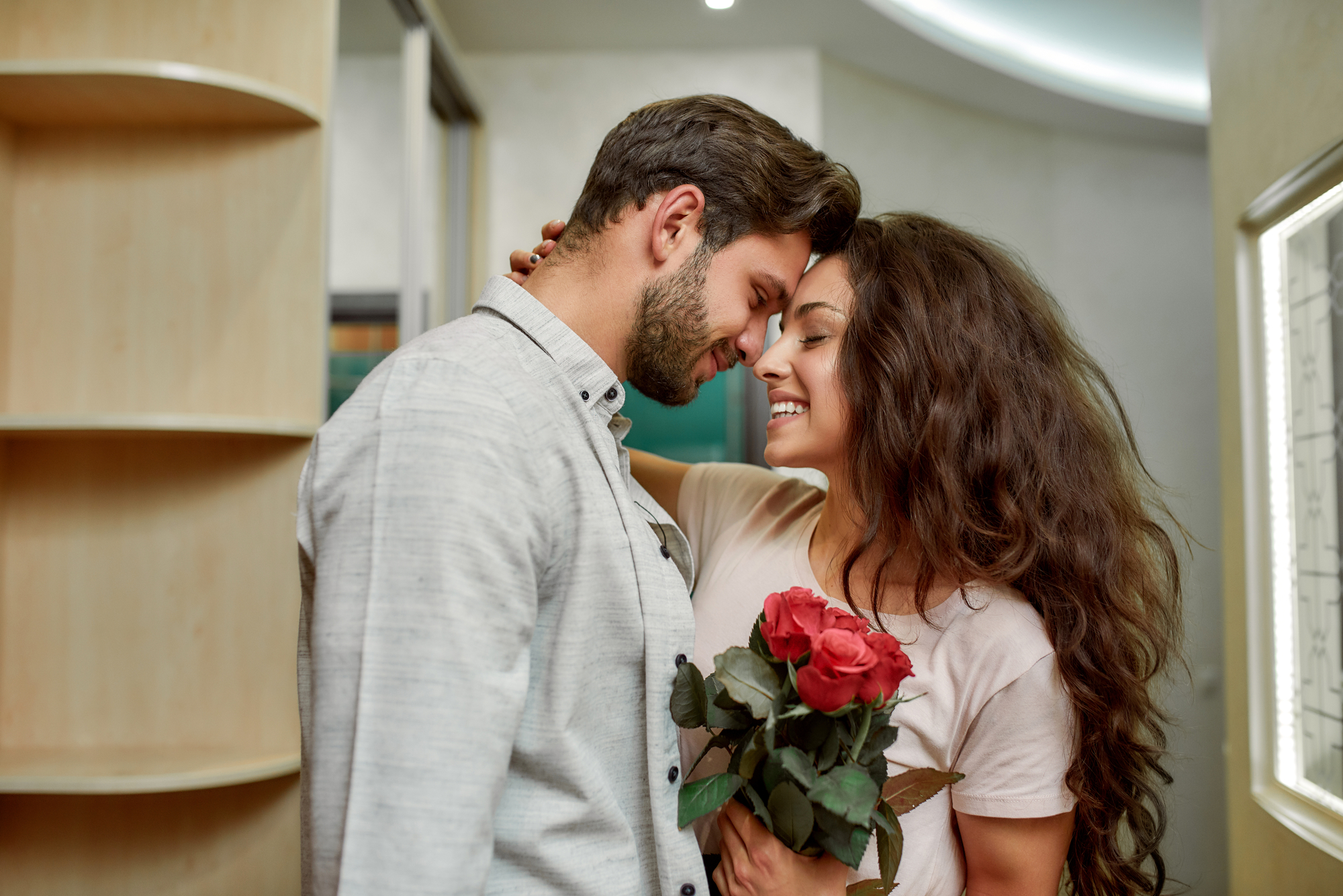 A couple stands close, forehead to forehead, smiling affectionately. The man, wearing a light button-up shirt, has his eyes closed. The woman, holding a bouquet of red roses, has long, wavy hair and appears to be laughing gently, looking at him.