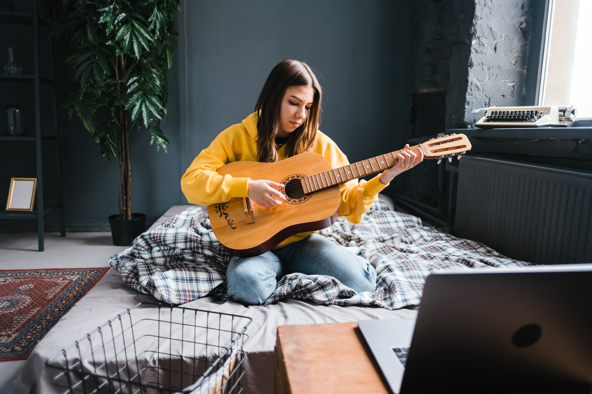A young woman with long brown hair sits cross-legged on a bed covered with a plaid blanket, playing an acoustic guitar. She wears a yellow hoodie and blue jeans. The room has gray walls, a large plant, and a laptop open on a desk in the foreground.