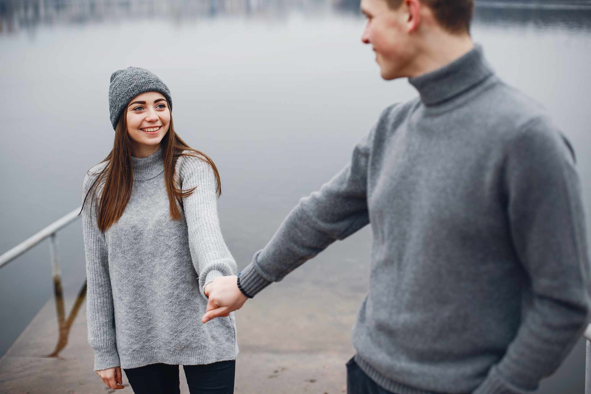 A young woman and a young man, both wearing gray sweaters, stand holding hands near a calm lake. The woman is smiling and wearing a gray beanie, while the man, slightly out of focus, looks at her with a smile. It’s a serene, overcast day.