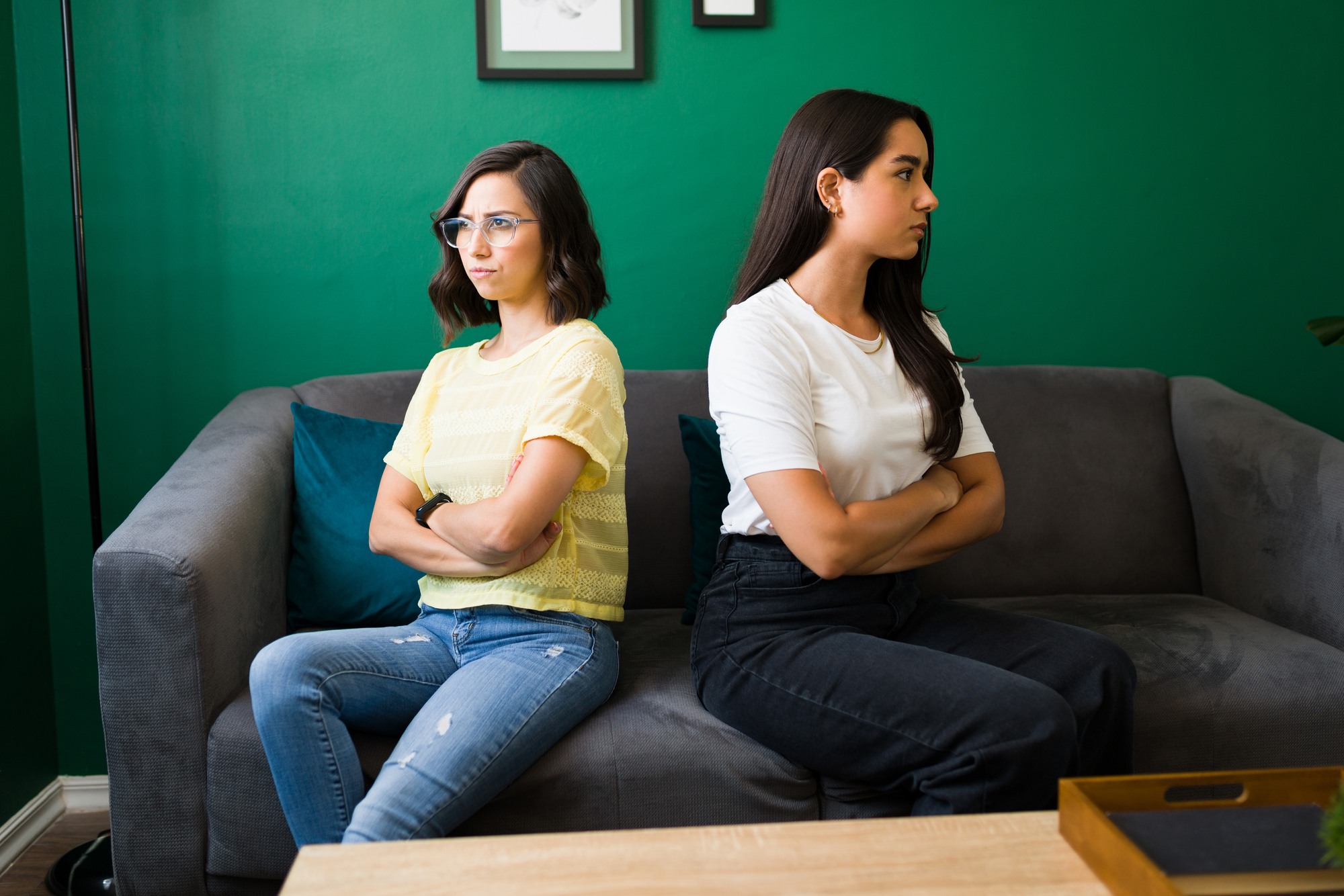 Two women are sitting on a gray couch with their arms crossed, facing away from each other. One wears a yellow shirt and glasses, the other wears a white shirt. The background features a green wall with framed pictures. They seem upset or in disagreement.
