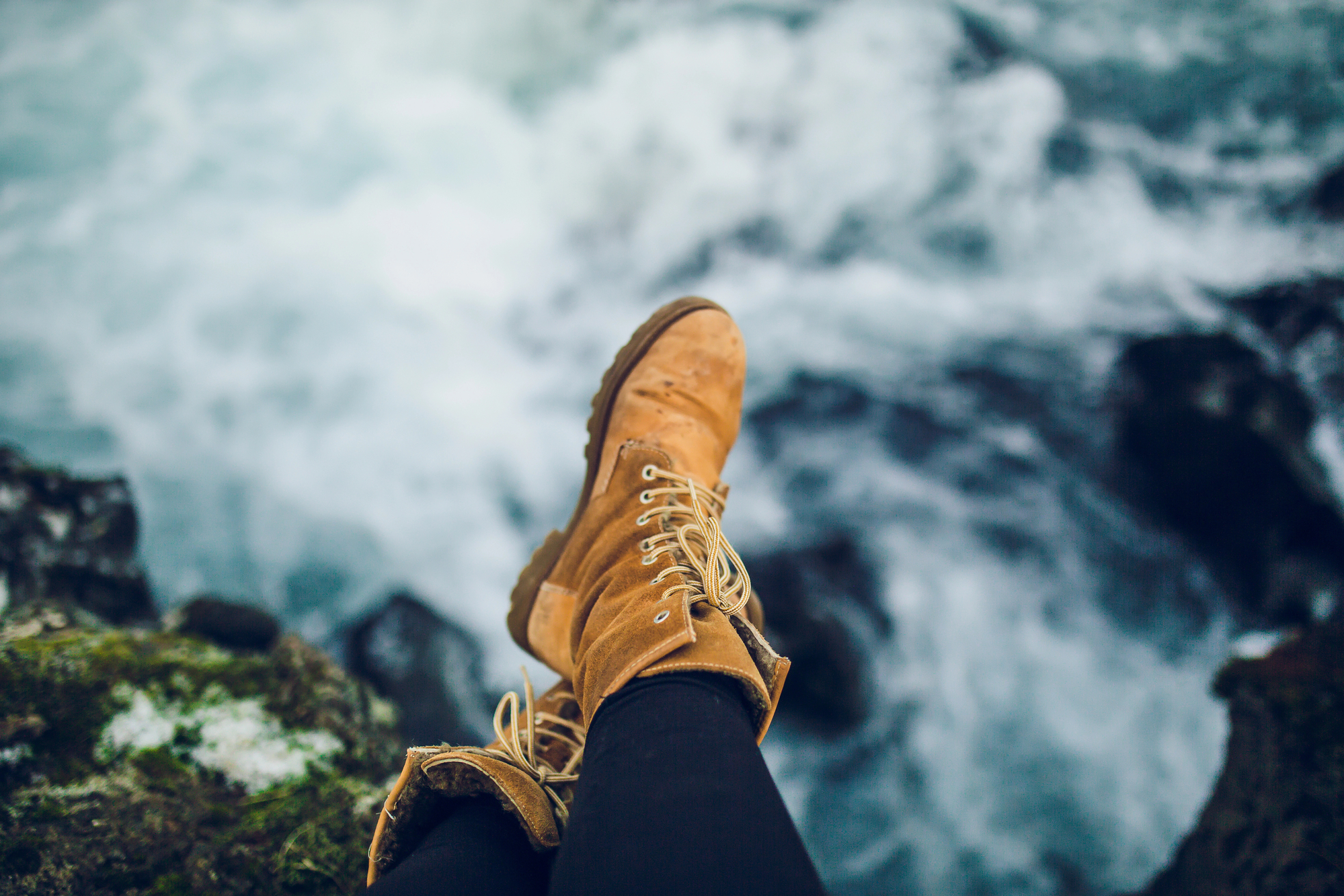 A pair of legs clad in black pants, with brown leather boots, are seen dangling over a rocky edge. Below them, foamy waves crash against rocks, creating a dramatic and dynamic water scene. The overall ambiance is rugged and adventurous.