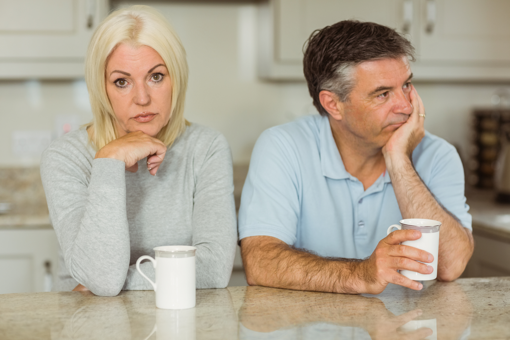 A woman with blonde hair and a serious expression sits at a kitchen counter, resting her chin on her hand. A man with graying hair sits next to her, looking pensive with his hand supporting his face. Both have coffee mugs in front of them.