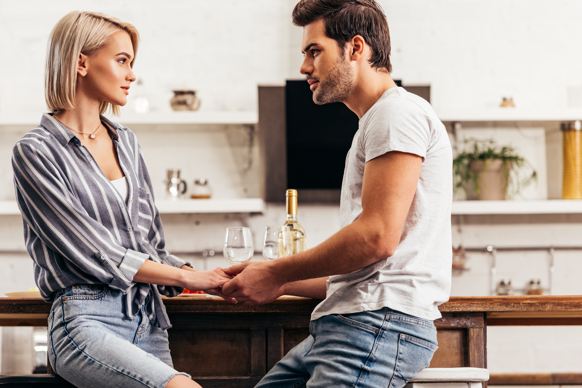 A man and a woman sit on stools in a modern kitchen while holding hands and looking at each other. The woman has blonde hair and is wearing a striped shirt and jeans. The man has dark hair and is wearing a white t-shirt and jeans. A bottle of wine and glasses are on the counter.