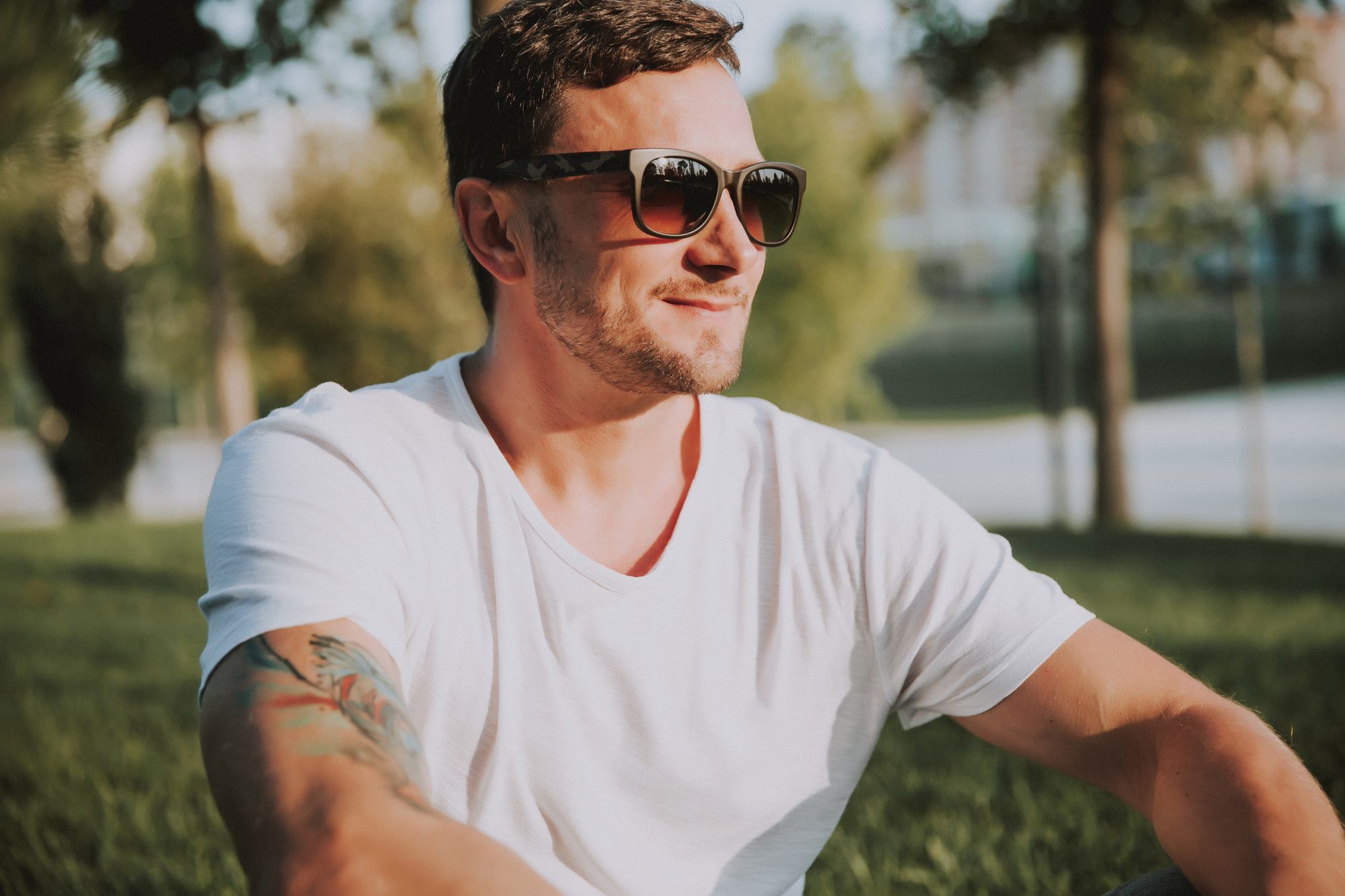 A man with short hair, wearing sunglasses and a white T-shirt, sits on the grass in a sunlit park. He has a tattoo on his right forearm and is smiling while looking to the side. Trees and a walkway are visible in the background.